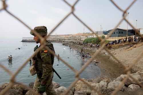 A member of the Spanish military keeps watch by a fence, as people waddle through water attempting to enter Spain, and in the background people who just entered Spain walk back to return to Morocco