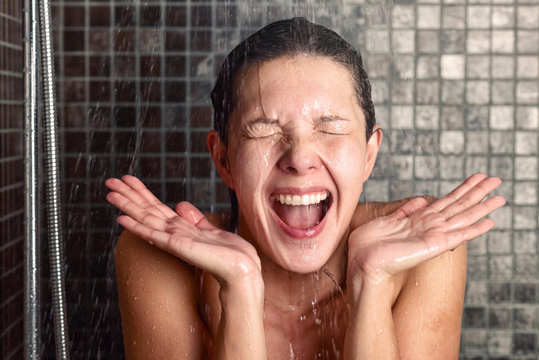Young woman reacting in shock to hot or cold shower water as she stands under the shower head washing her hair eyes closed with