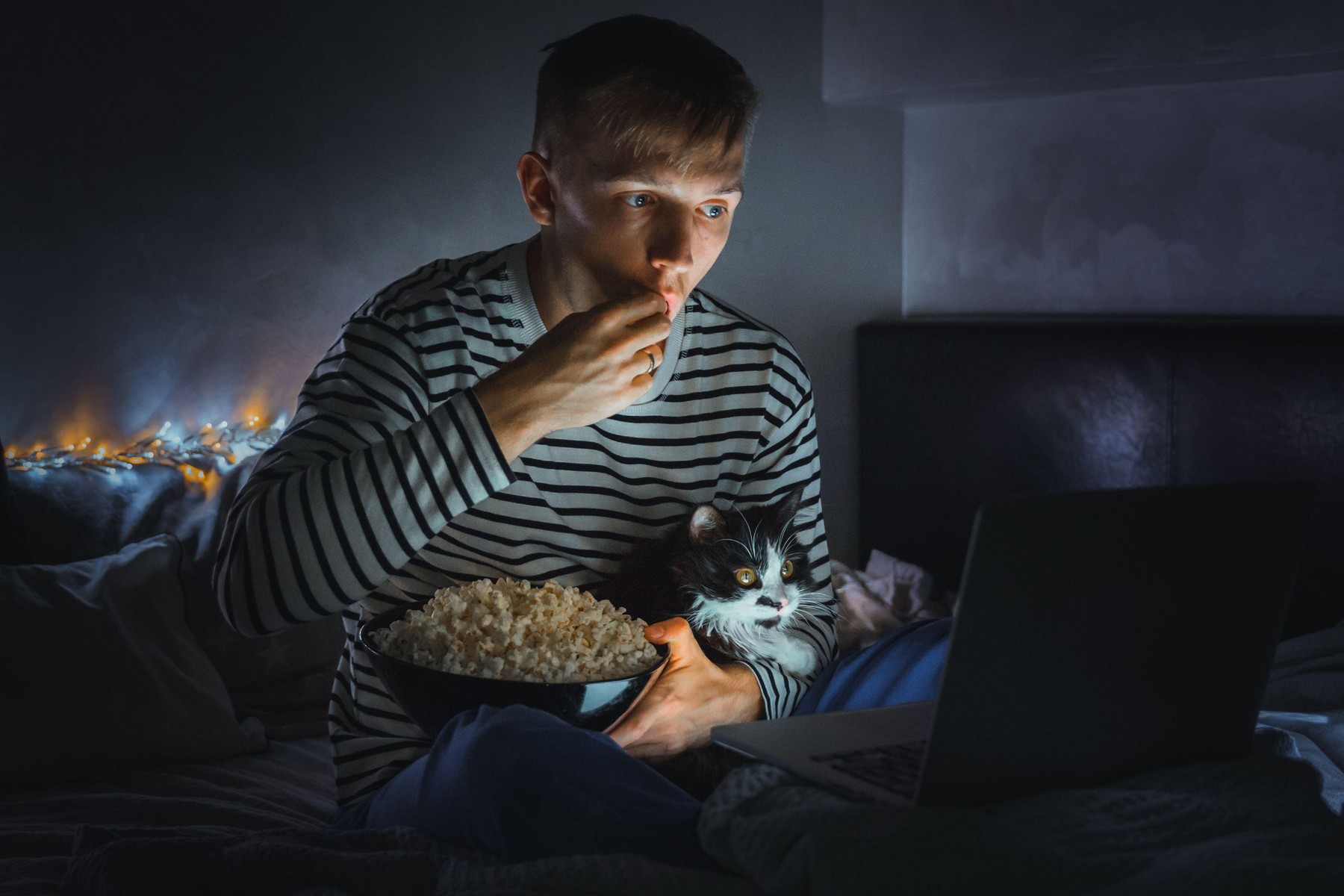 young man with black cat watching a movie eating popcorn on TV at home. Movie night. Relax,rest watching a horror film or video on screen. Background