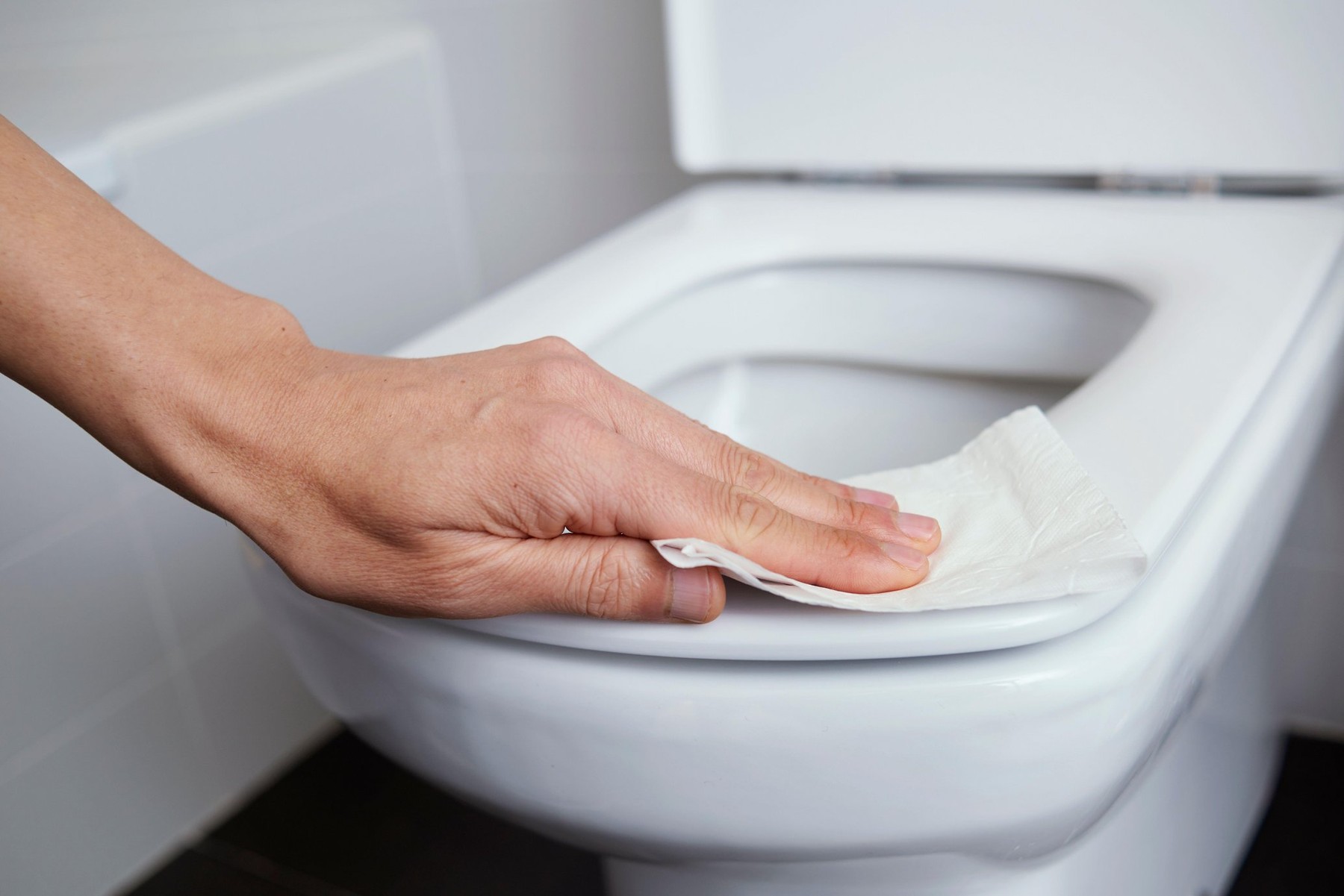 closeup of a caucasian man cleaning the toilet seat with a piece of toilet paper in a tiled bathroom