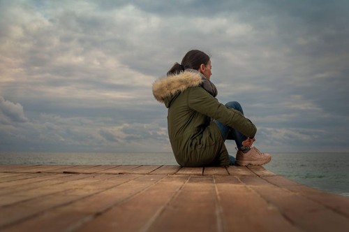 Woman wearing a parka coat sitting by the sea, stormy sky, lonelyness concept