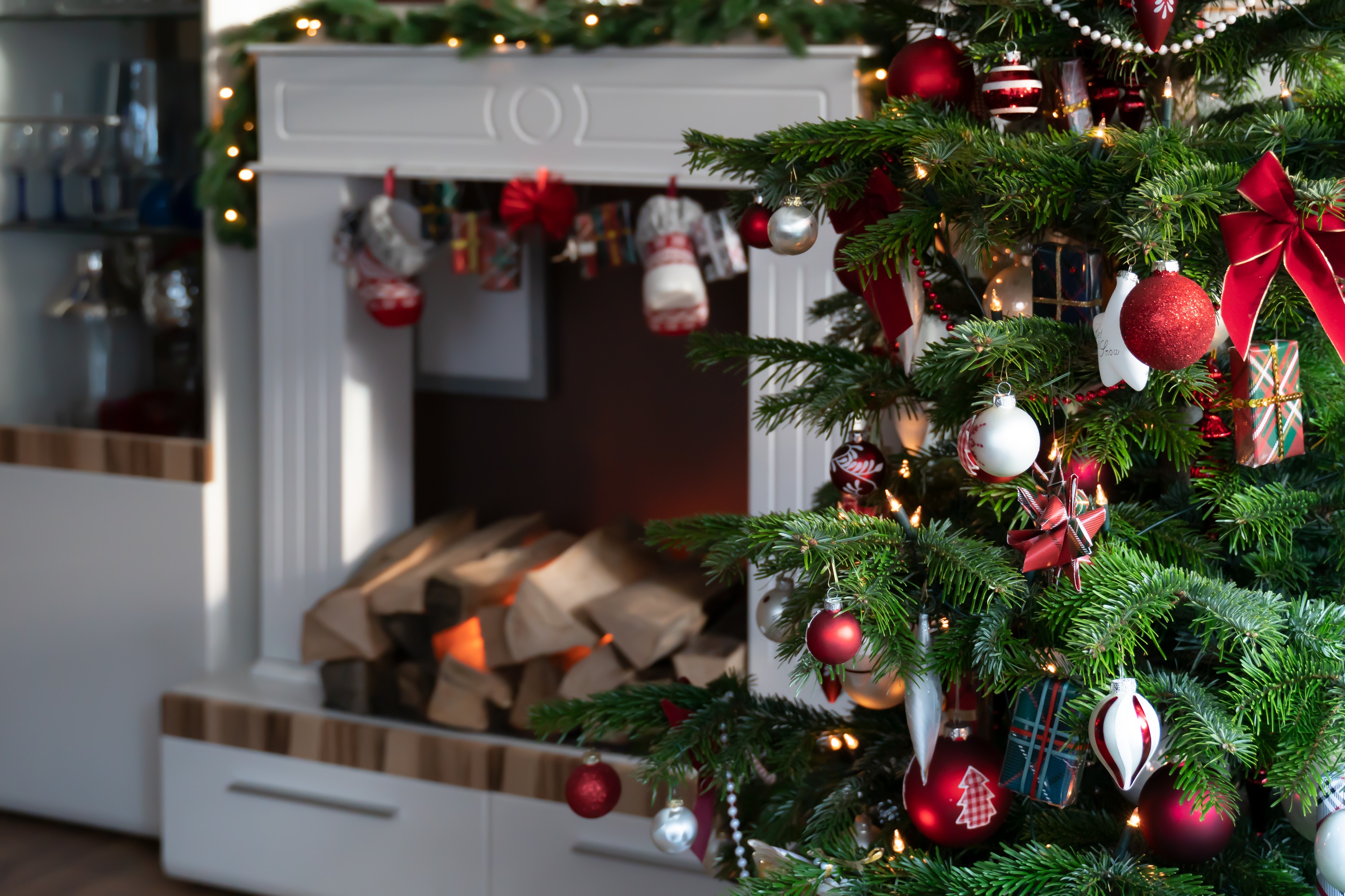 Christmas tree is in the living room is decorated in red and white. In the background is electric fireplace in white cabinet.