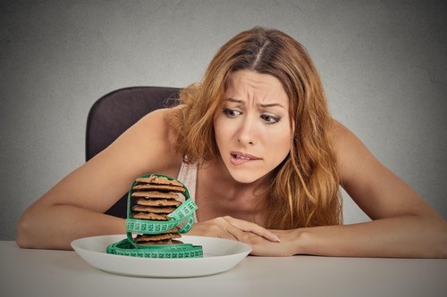 Portrait young unhappy woman craving sugar sweet cookies but worried about weight gain sitting at table isolated grey wall background. Human face expr