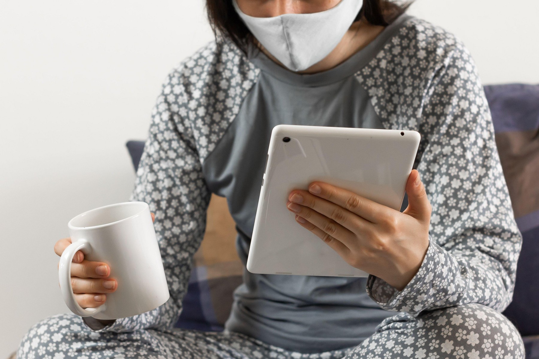 Young woman in protective mask to prevent coronavirus sits in a bed and works with a tablet. Home office concept. Freelancer in nightwear works online