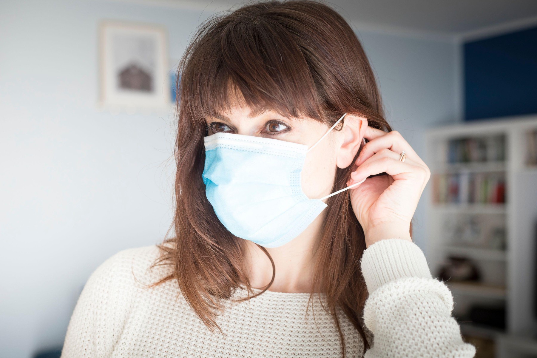 Caucasian attractive woman with long hair and bangs, putting on a surgical blue face mask during covid-19 or coronavirus pandemic.