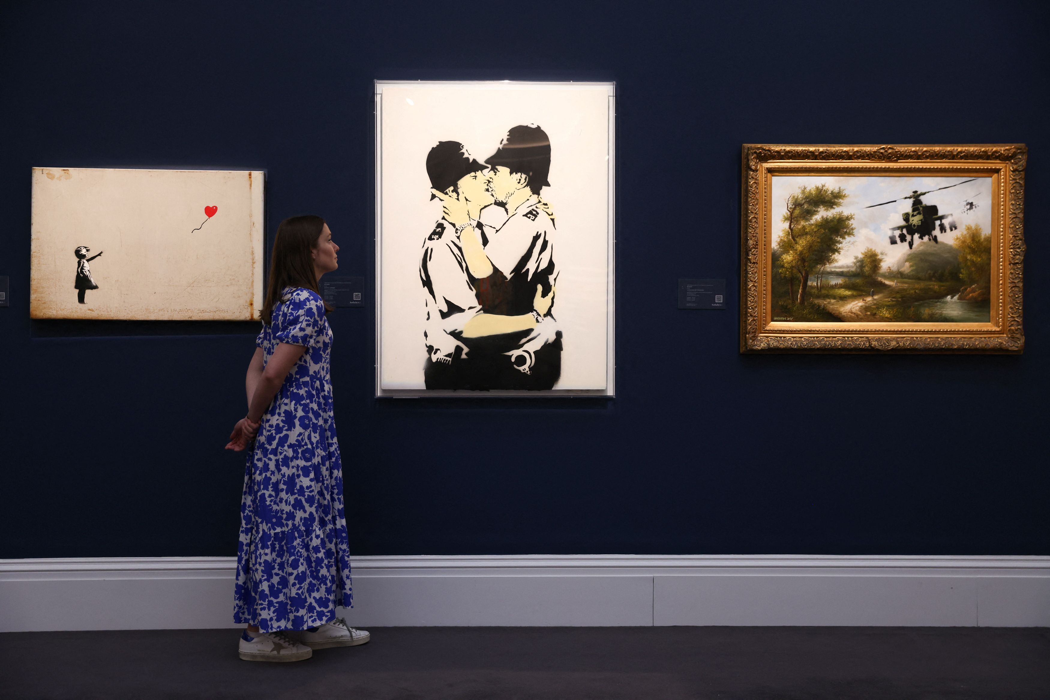 A Sotheby's employee poses with three artworks from the Robbie Williams Collection, including 'Kissing Coppers, 2005' by Banksy, on display ahead of auction at Sotheby's in London