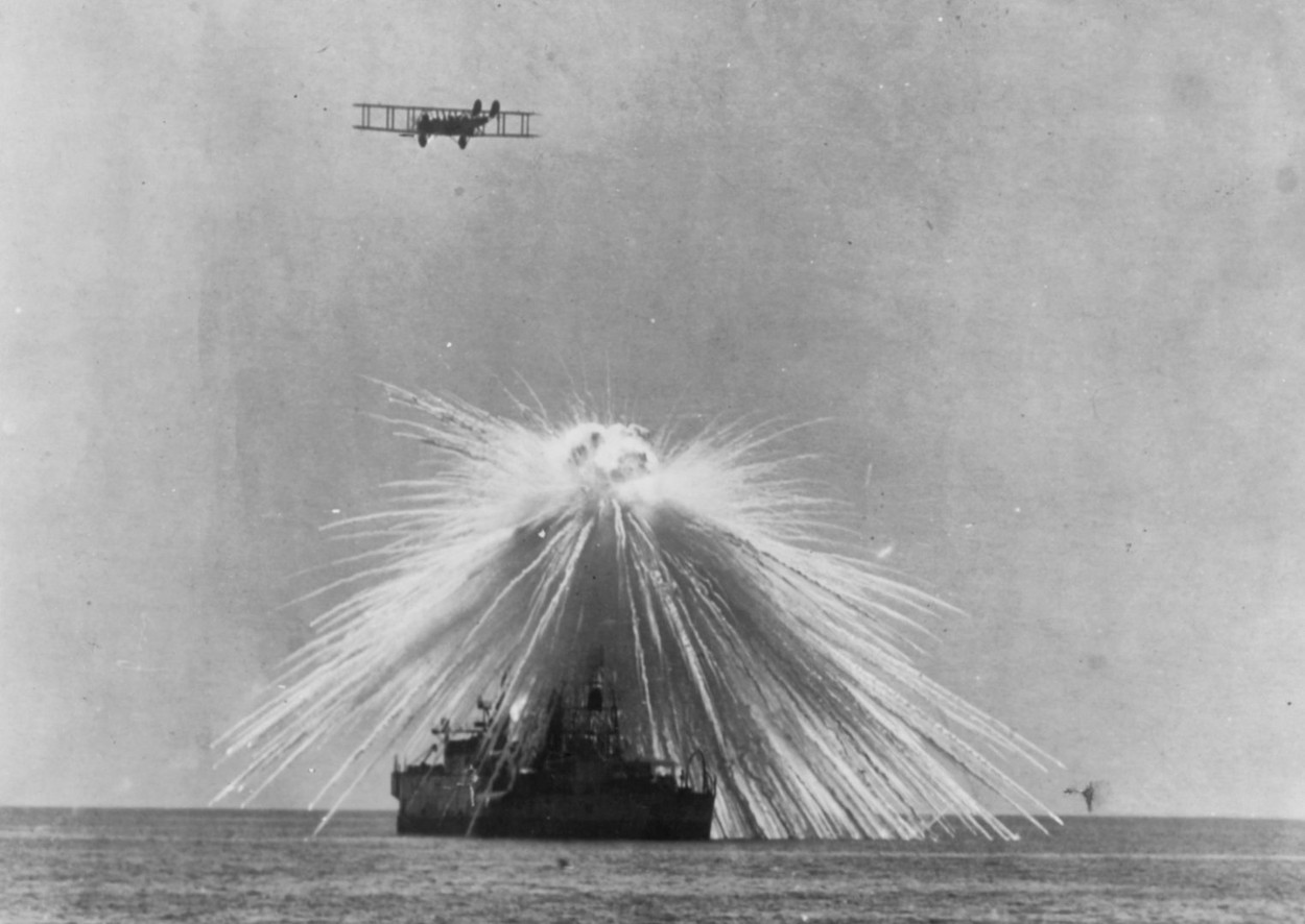 Ex-USS Alabama (BB-8) - A white phosphorus bomb explodes on a mast top, while the ship in use as a target in Chesapeake Bay, 23 September 1921. An Army Martin twin-engine bomber is flying overhead. 23 September 1921