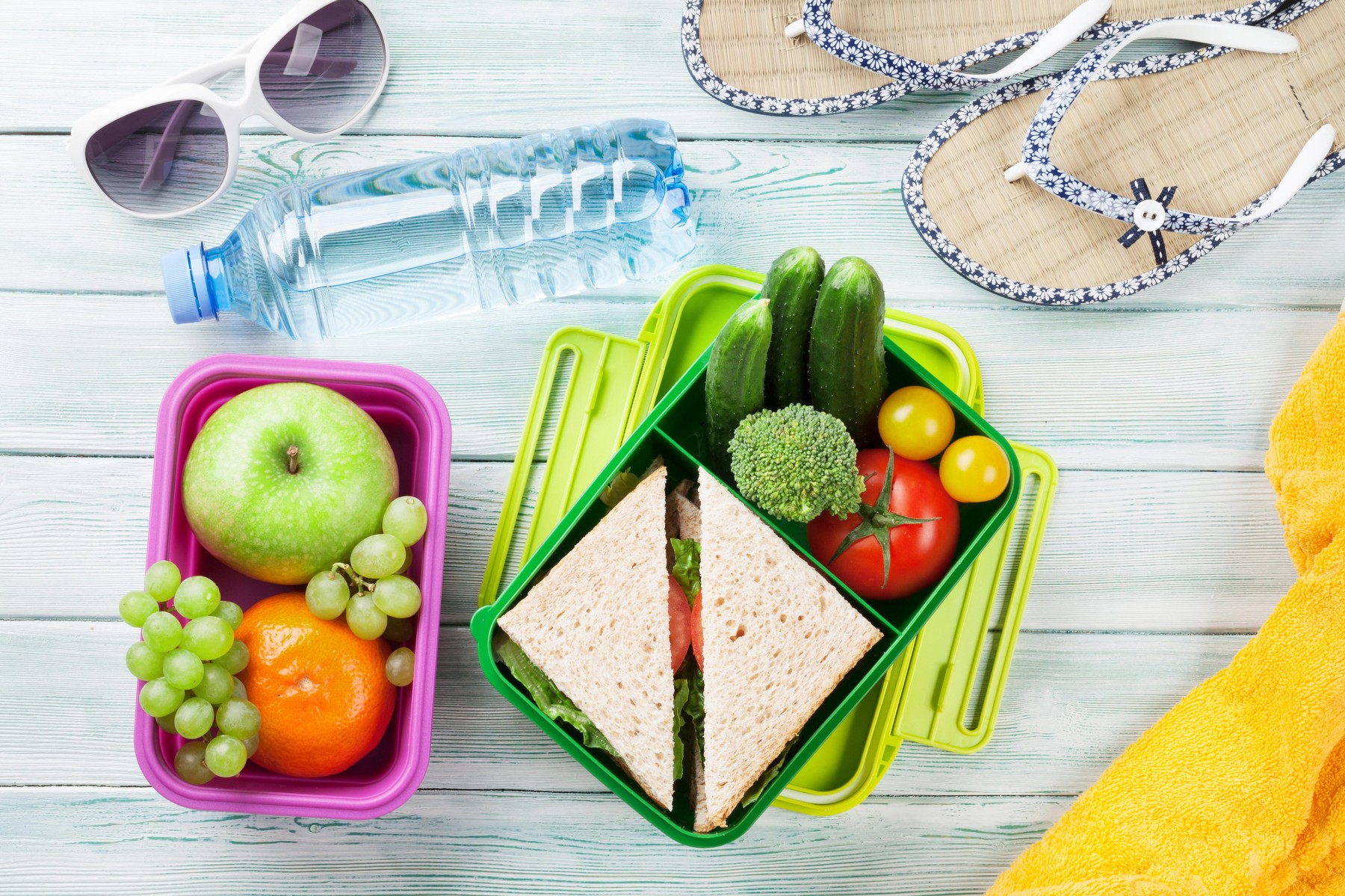 Lunch box with vegetables and sandwich on wooden background. Beach take away food box, towel and glasses. Top view