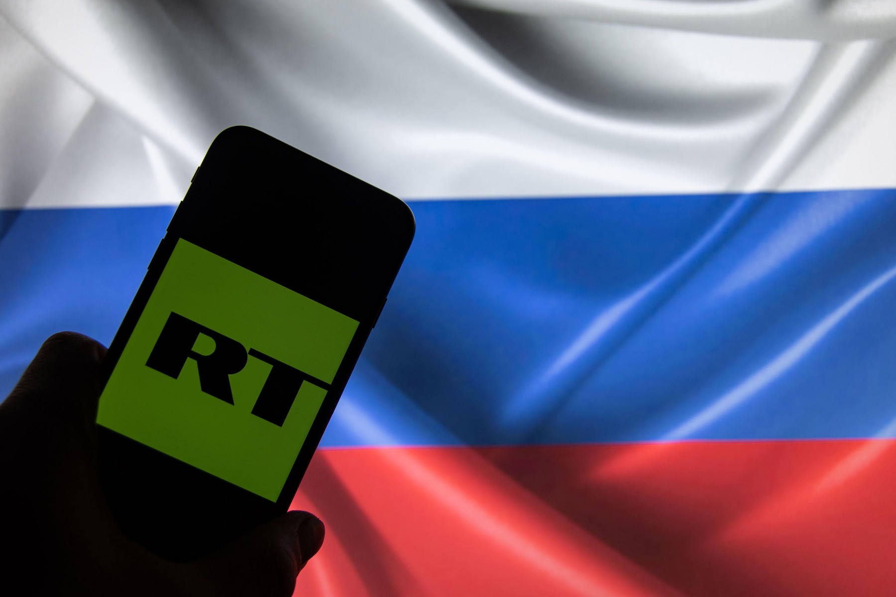 Rheinbach, Germany 4 March 2022, The "Russia Today" brand logo on the display of a smartphone in front of a Russian flag (focus on the brand logo)