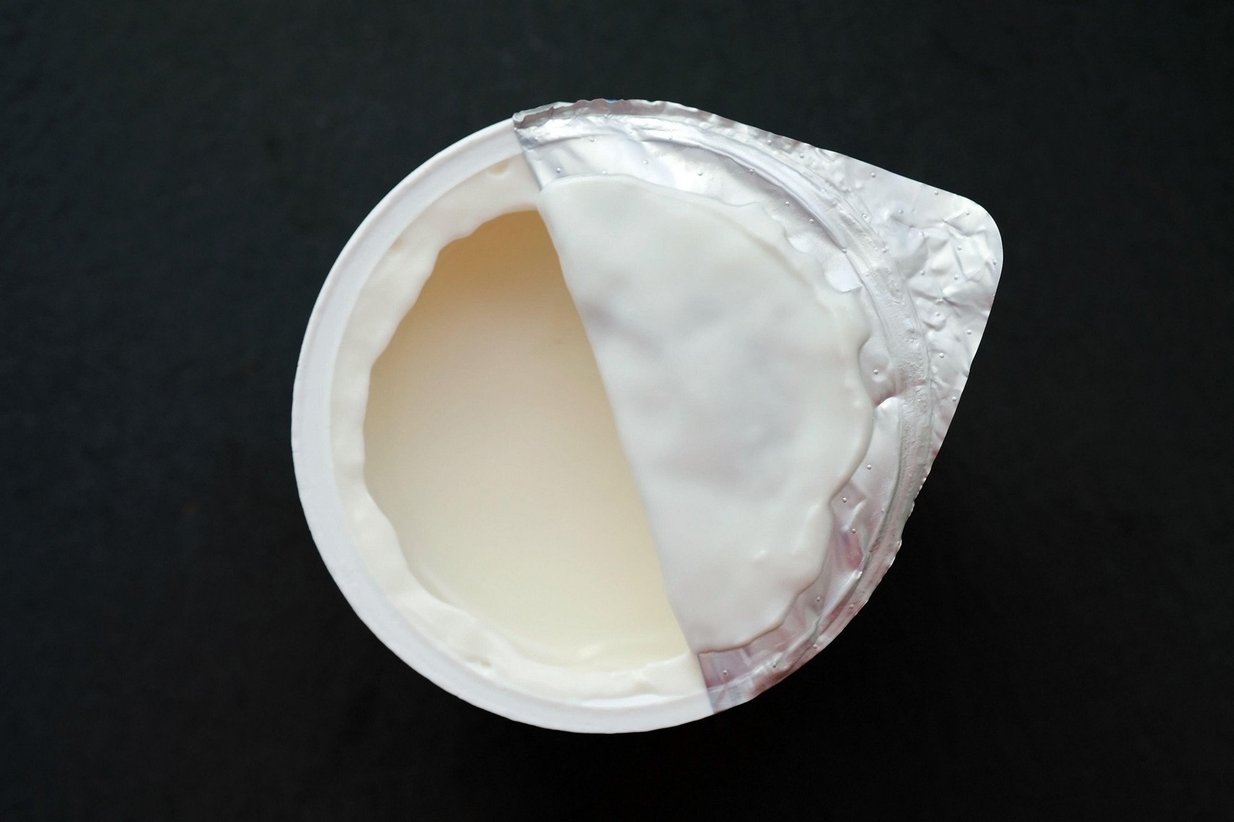White yogurt in a plastic cup against a black background. Children's yogurt is half-open from above.