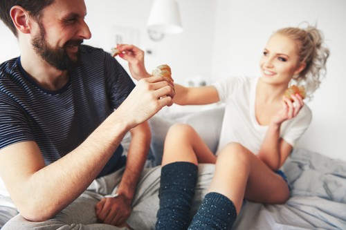 Couple sitting on bed, fooling around, woman sharing pastry with man