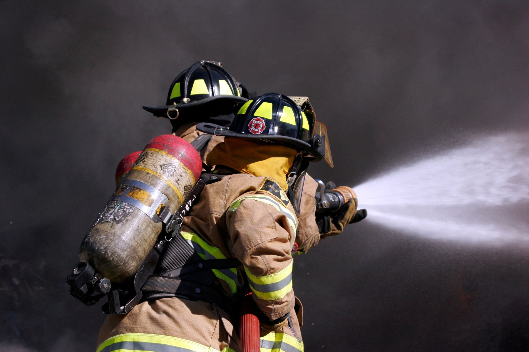 Two firefighters using a hoseline to extinguish a car fire while being surrounded by smoke and spray