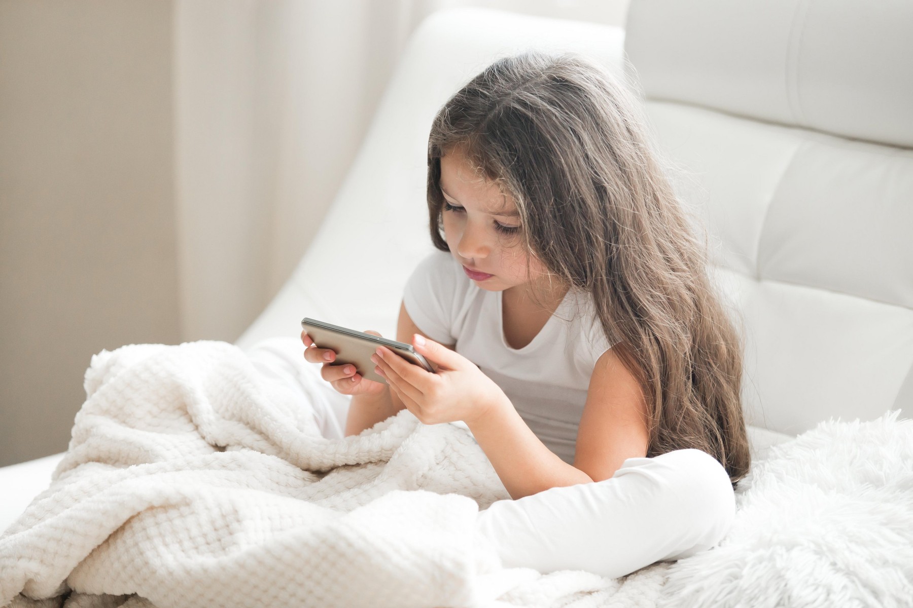 Little girl playing games or using app on tablet sitting on white coach in living room. Beautiful kid girl sitting on cozy