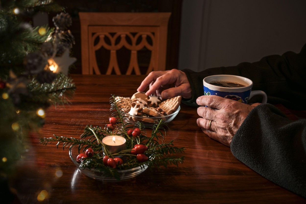 Hands of an elderly single man sitting alone at a table with Christmas cookies, coffee and festive decoration next to an empty chair, lonely holidays