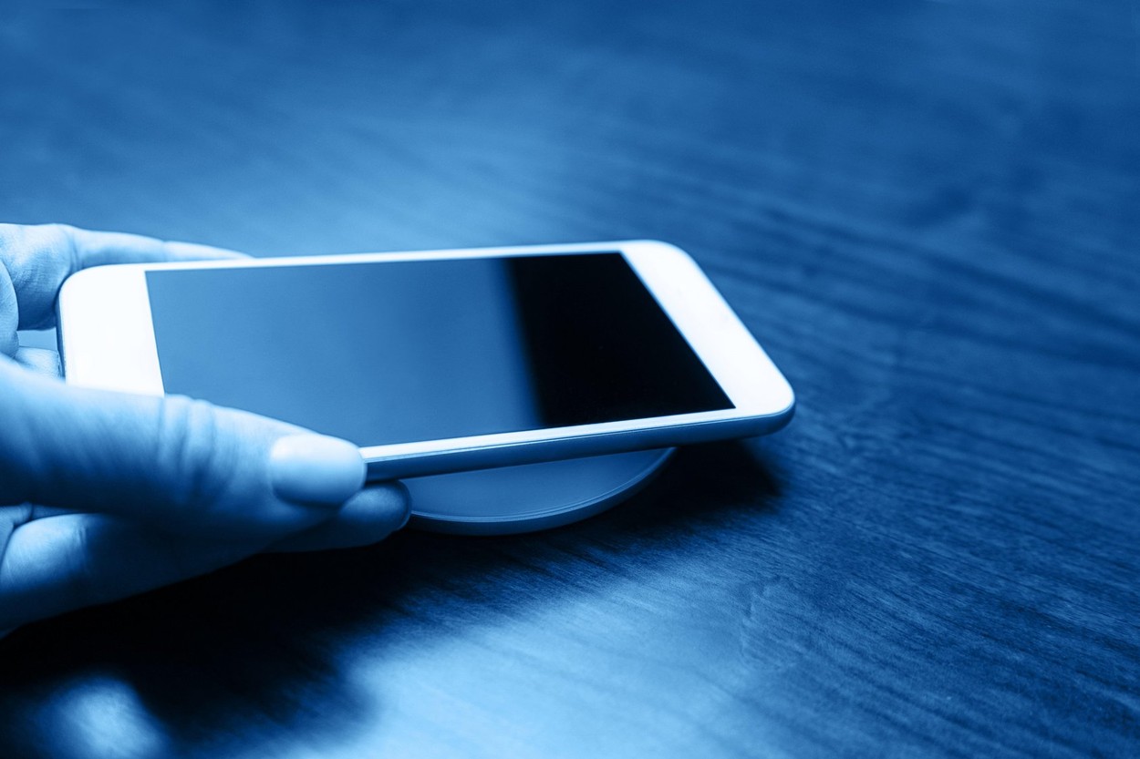 Hand holds a mobile smart phone for charging wirelessly by sharing battery via device in classic blue color. Charging technology. Tech Device Charge Sharing. Close up.