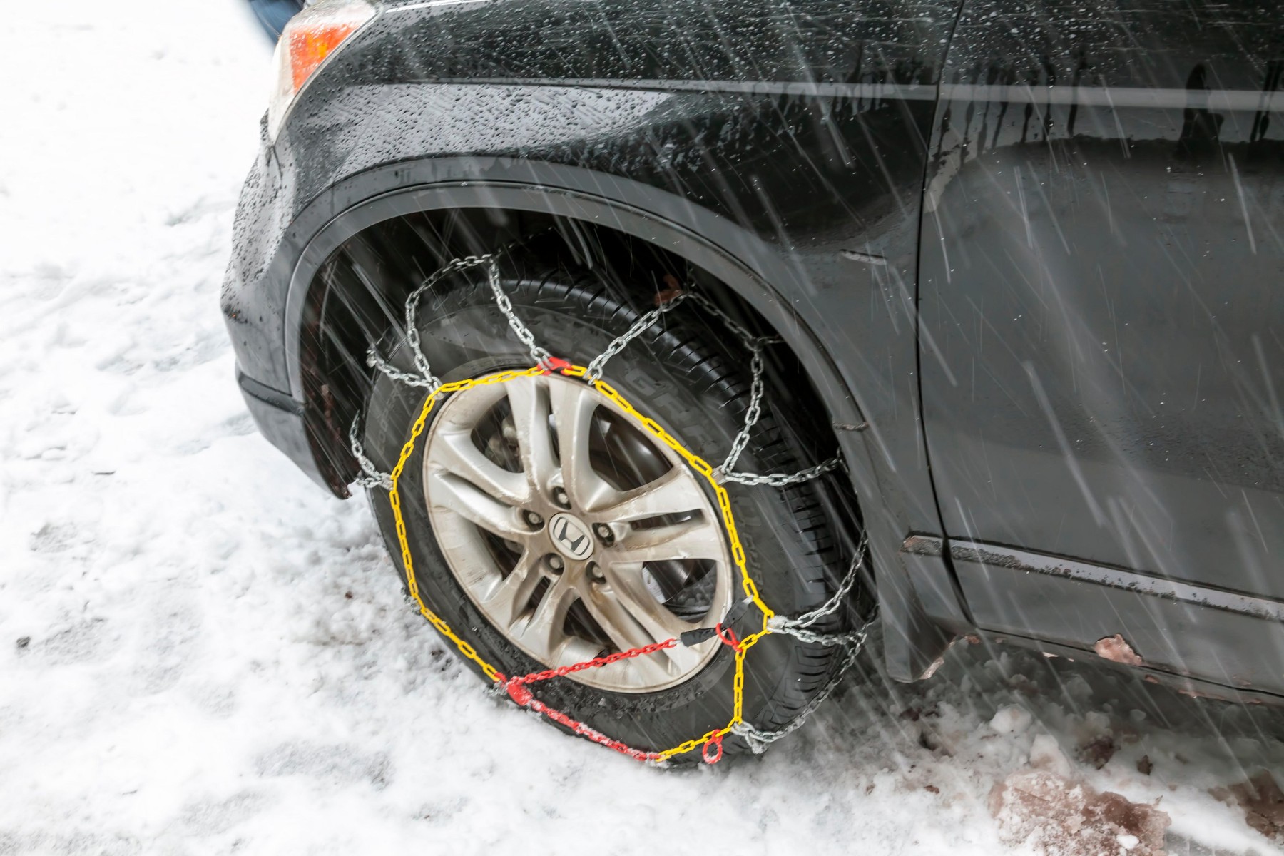 Snow chains installed on front wheel or tire of Honda passenger car on a snowy road while snowing.