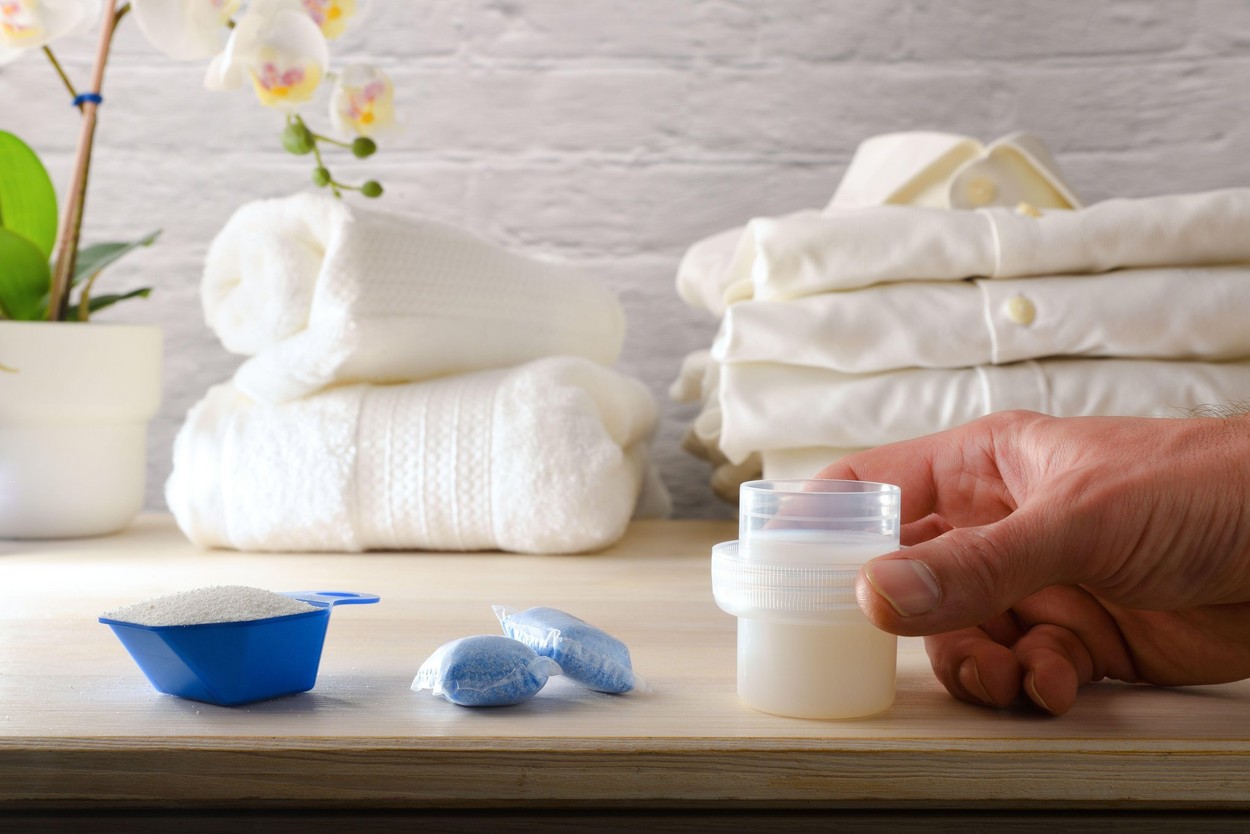 Hand showing different household laundry chemical products on wooden bench in a home laundry room with freshly washed white clothes in the background.