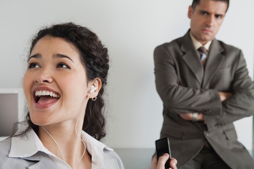 Woman distractedly listening to MP3 player at work, boss glaring in background