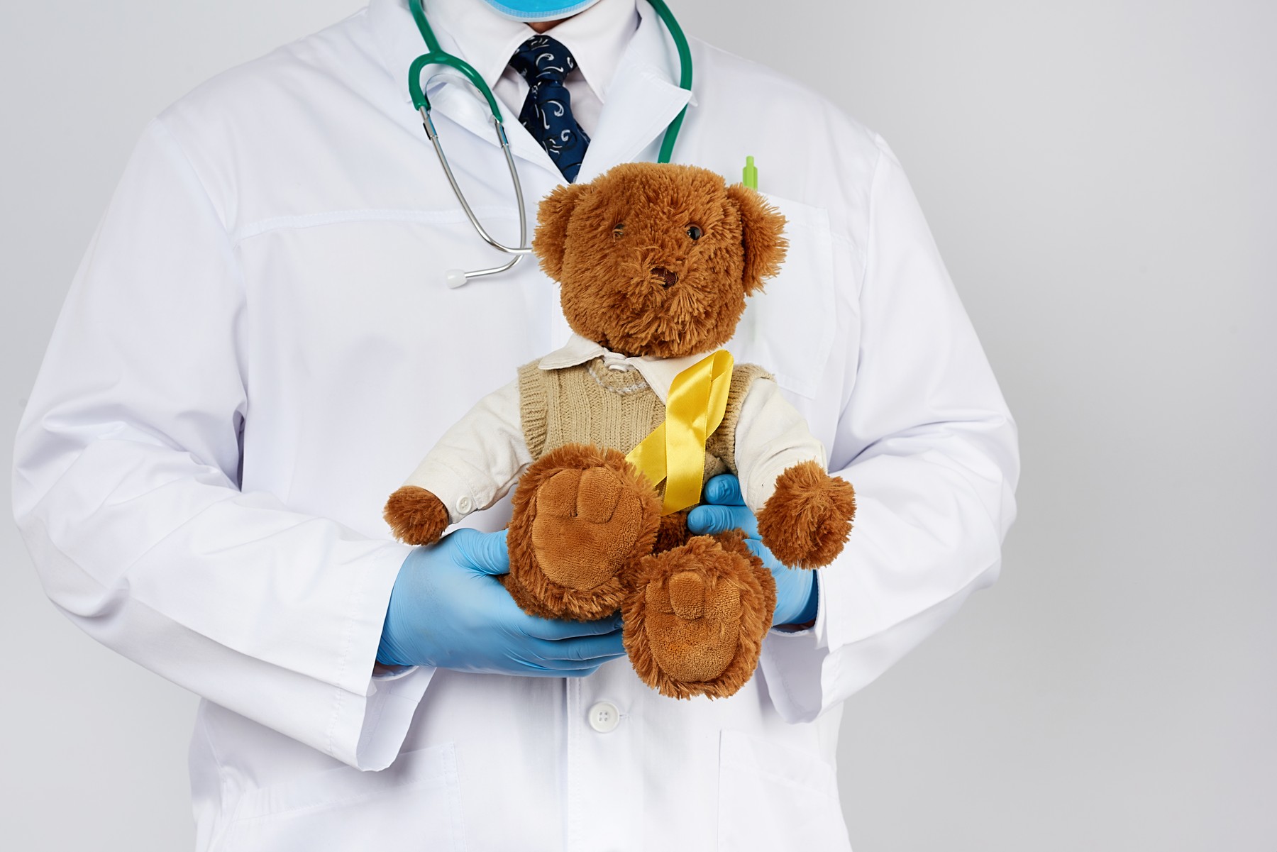 pediatrician in white coat, blue latex gloves holds a brown teddy bear with a yellow ribbon