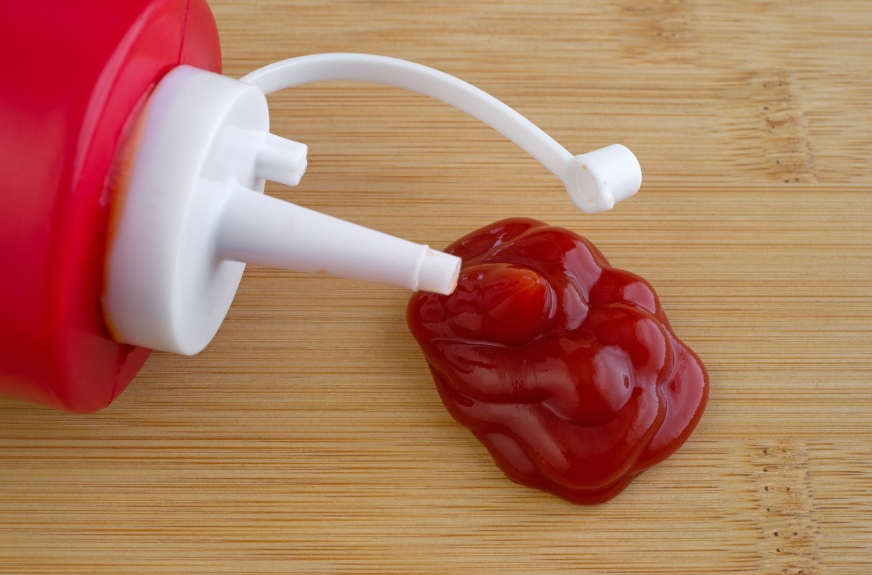 Close view of a bottle of ketchup on a wood cutting board with a blob under the spout.