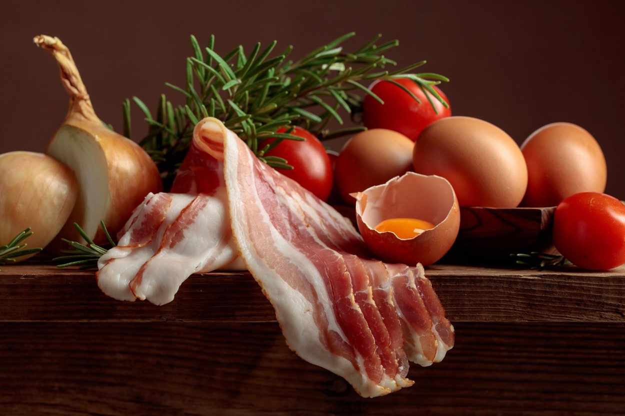 Smoked bacon with eggs, tomatoes, onions, and rosemary on an old wooden table.