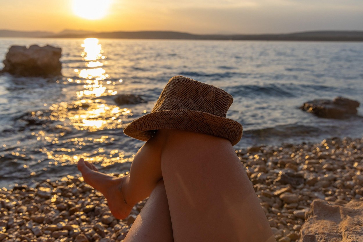 A barefoot girl on a beach with a hat on her knees, the Adriatic Sea in Croatia