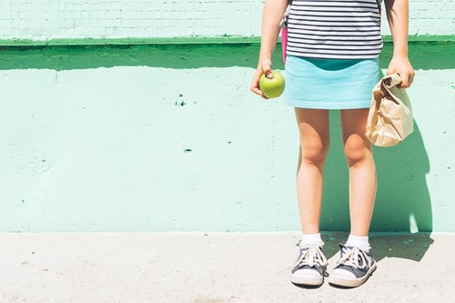 A little girl in sneakers is holding an apple and paper bag with a school snack against the background of a green wall.