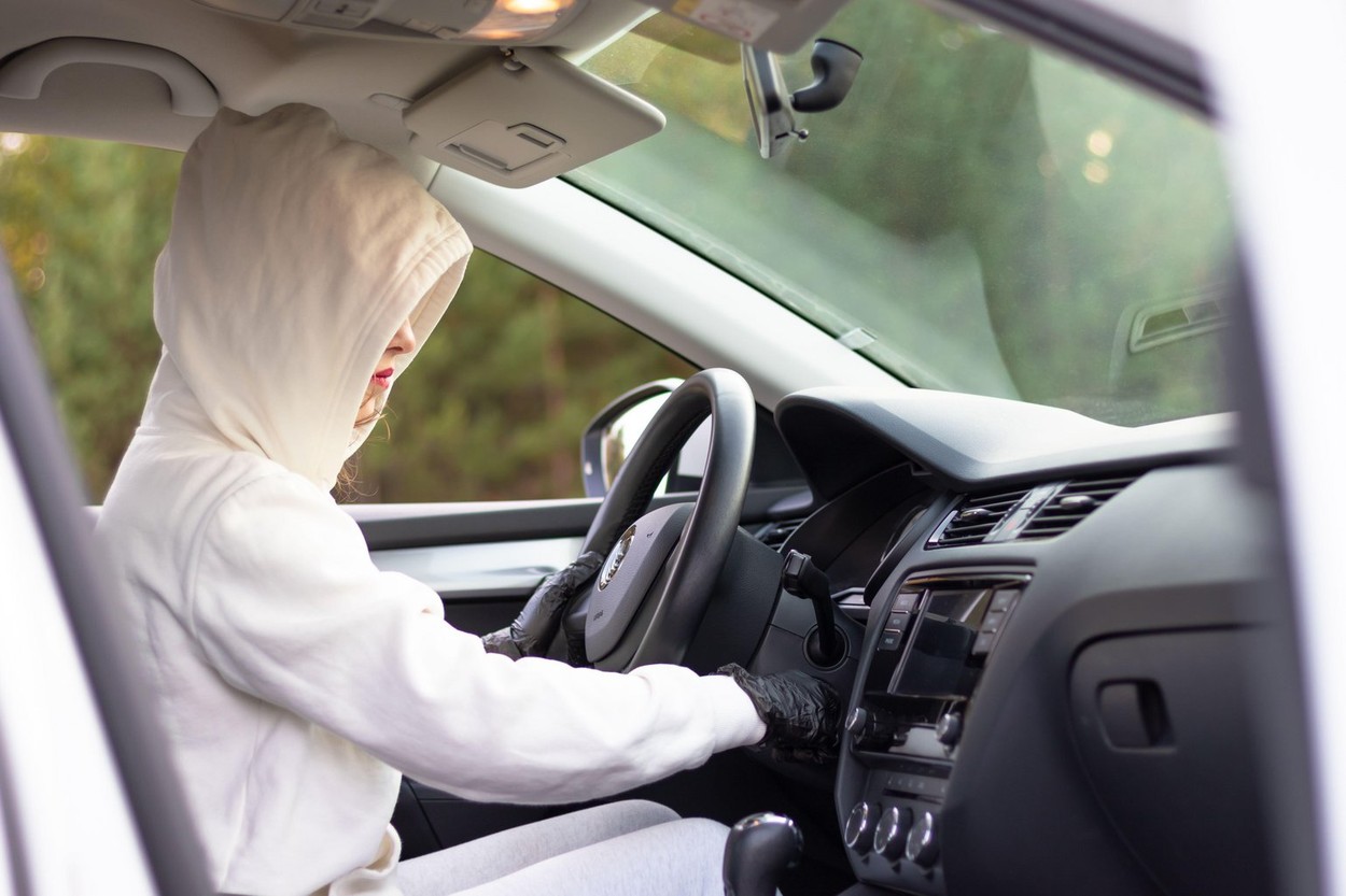 A beautiful young woman driver in a white jacket with a hood and black gloves sits at the wheel of a car. Selective focus. Portrait. Close-up