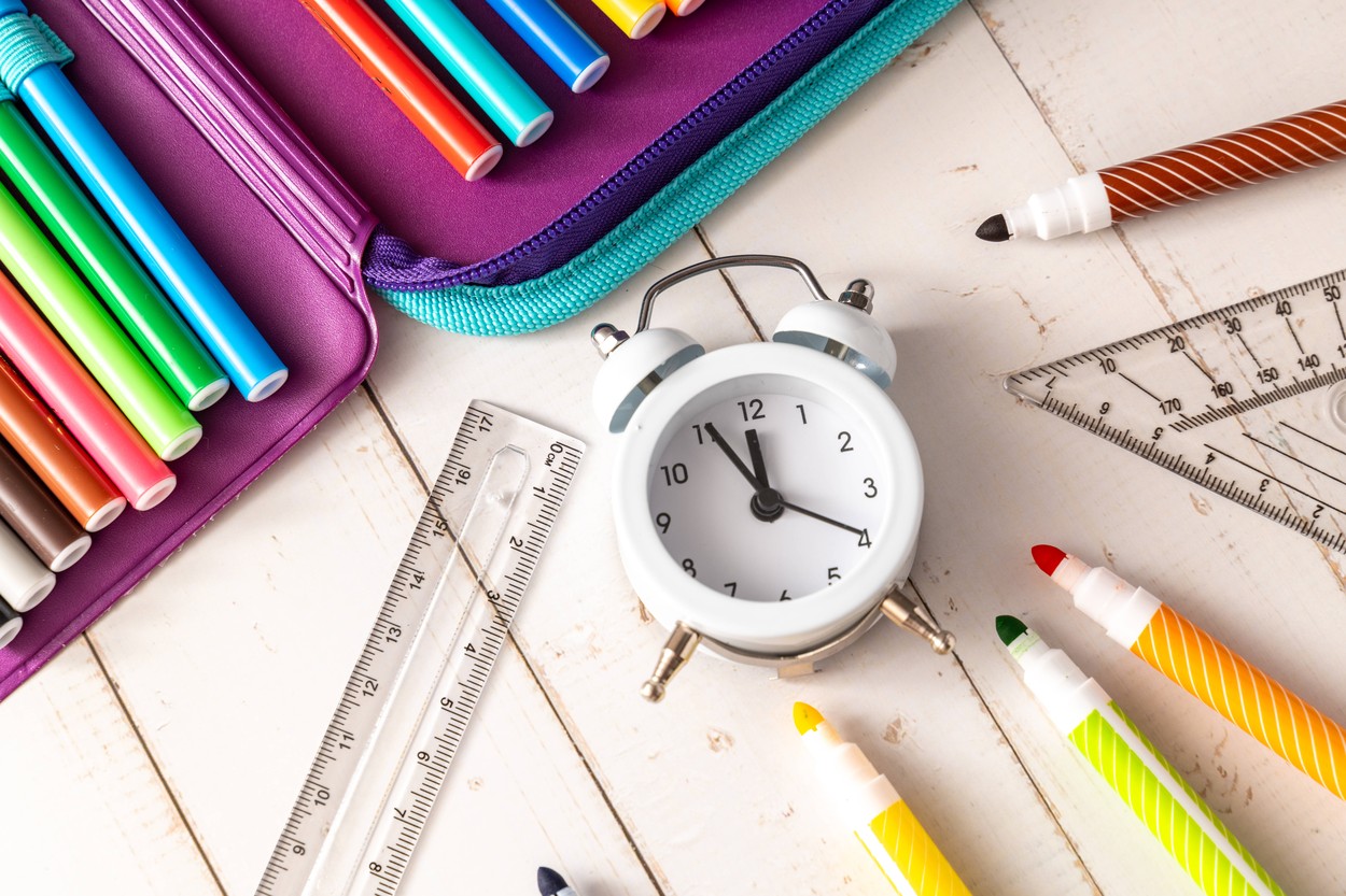 8 August 2023: Alarm clock with time 5 to 12 in front of school pencil case with colorful pens. school, vacations and ex