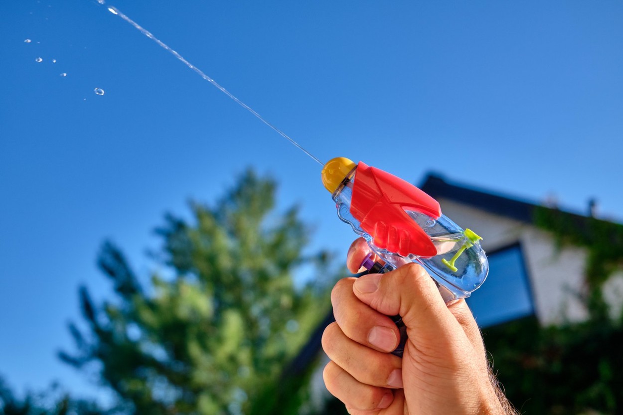 Colorful water pistol spraying out a jet of water in a caucasian male hand in front of  a house and the blue sky of a sunny summer day