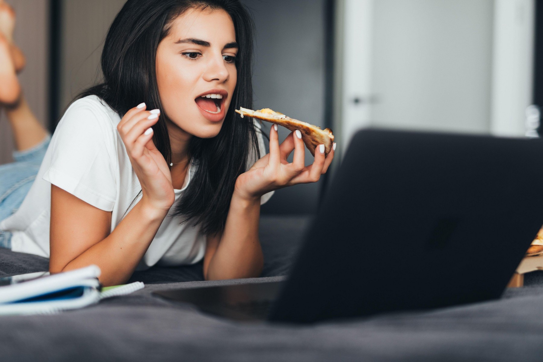 Young woman lying on a bed in bed at home while eating pizza and using laptop