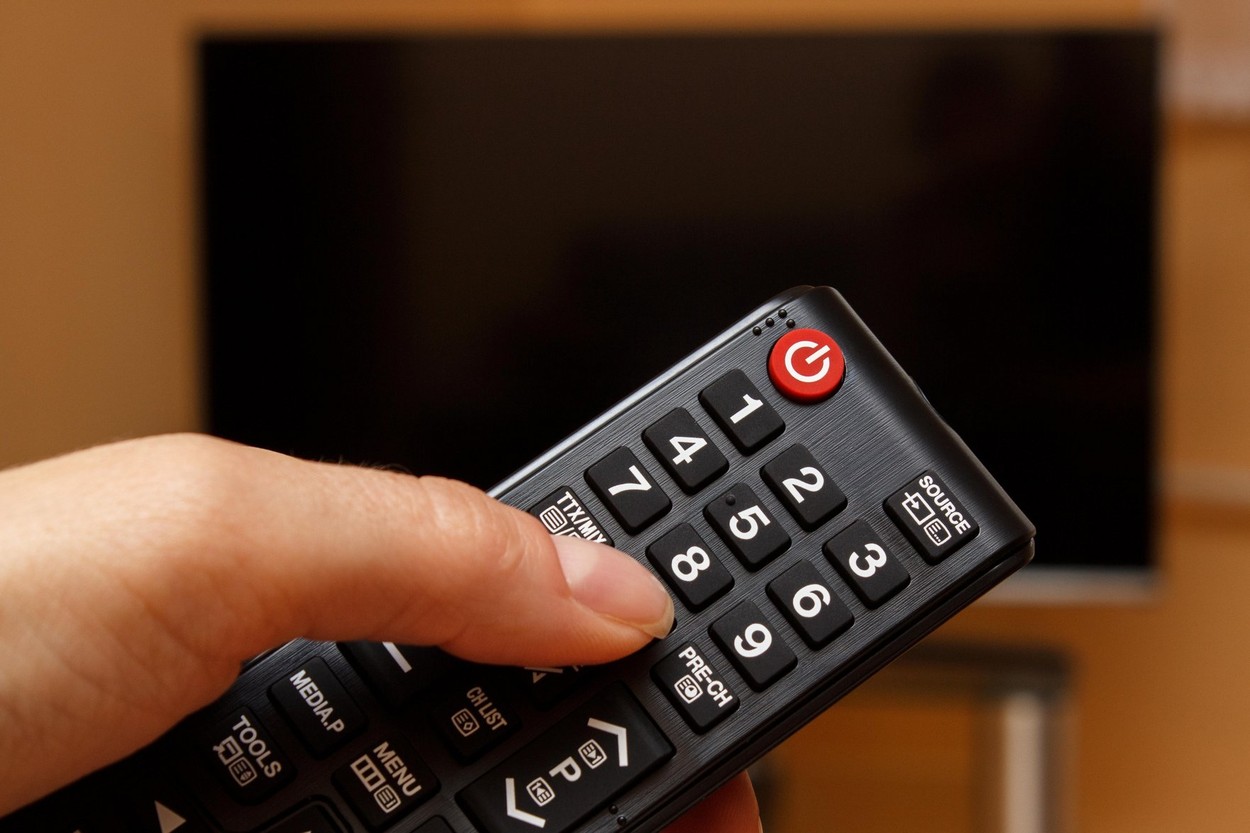 Hand holding infrared remote control for television and TV screen in background, choosing program to watch