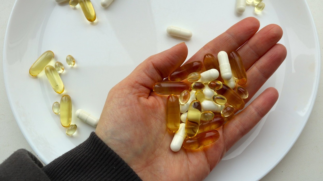 a woman's hand holds a handful of capsules of different colors and sizes over a large white plate with scattered pills