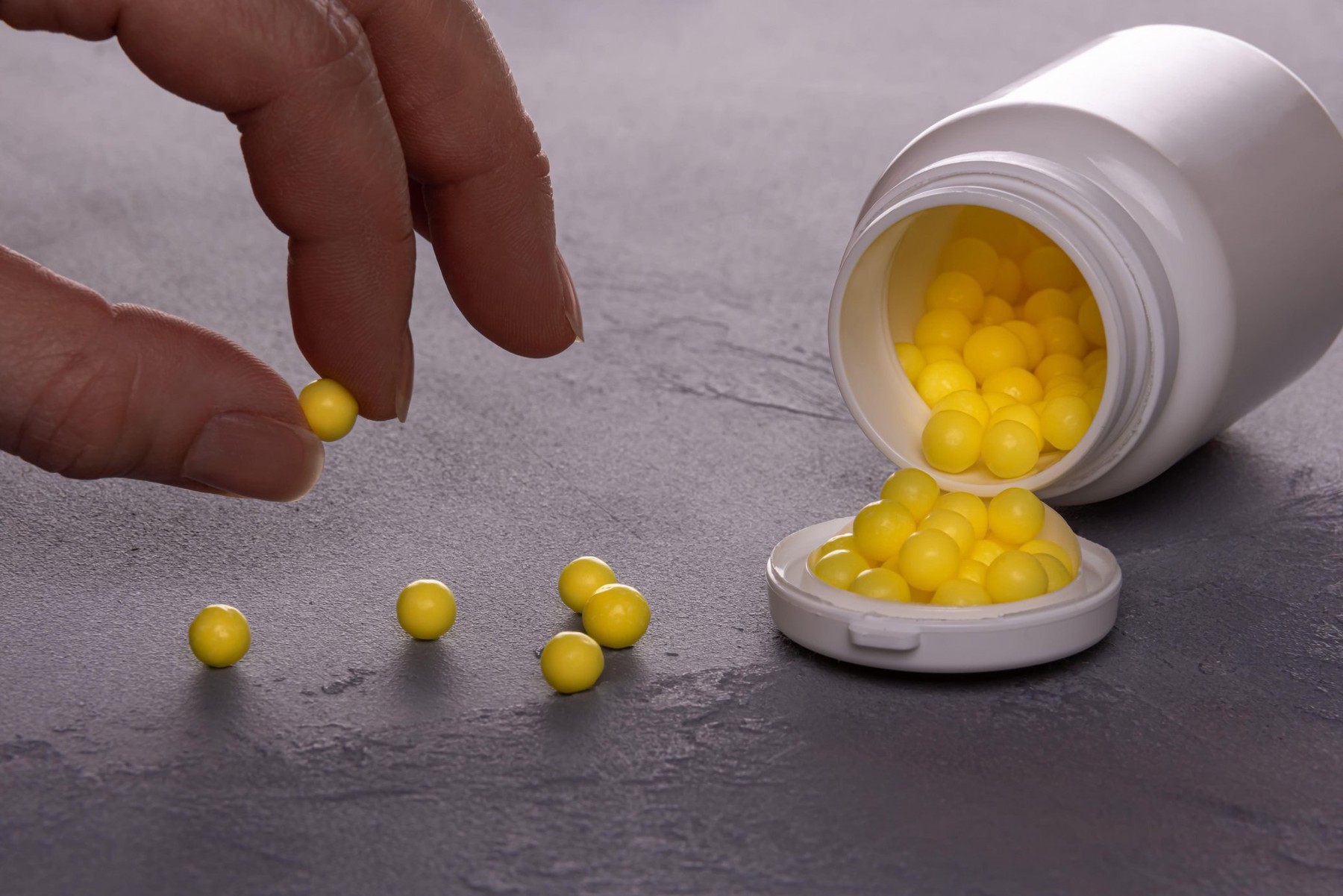 Close-up of opened white plastic medicine bottle lying on side and full of small round yellow pills of Vitamin C. Few pills are scattered over surface