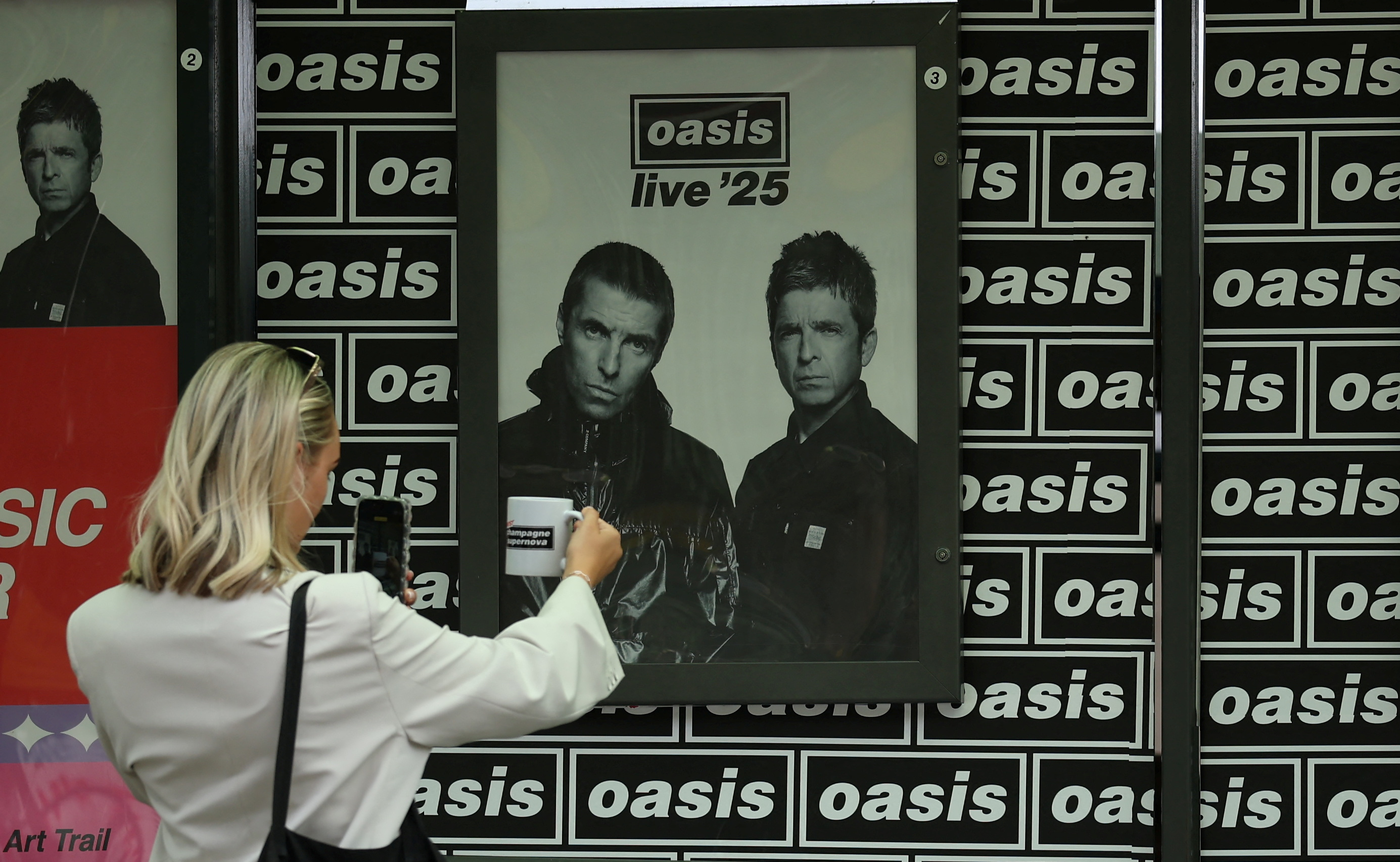 A woman takes photographs of a mug in front of Oasis posters ahead of their concerts in Heaton Park in Manchester, Britain, July 9, 2025. REUTERS/Phil Noble