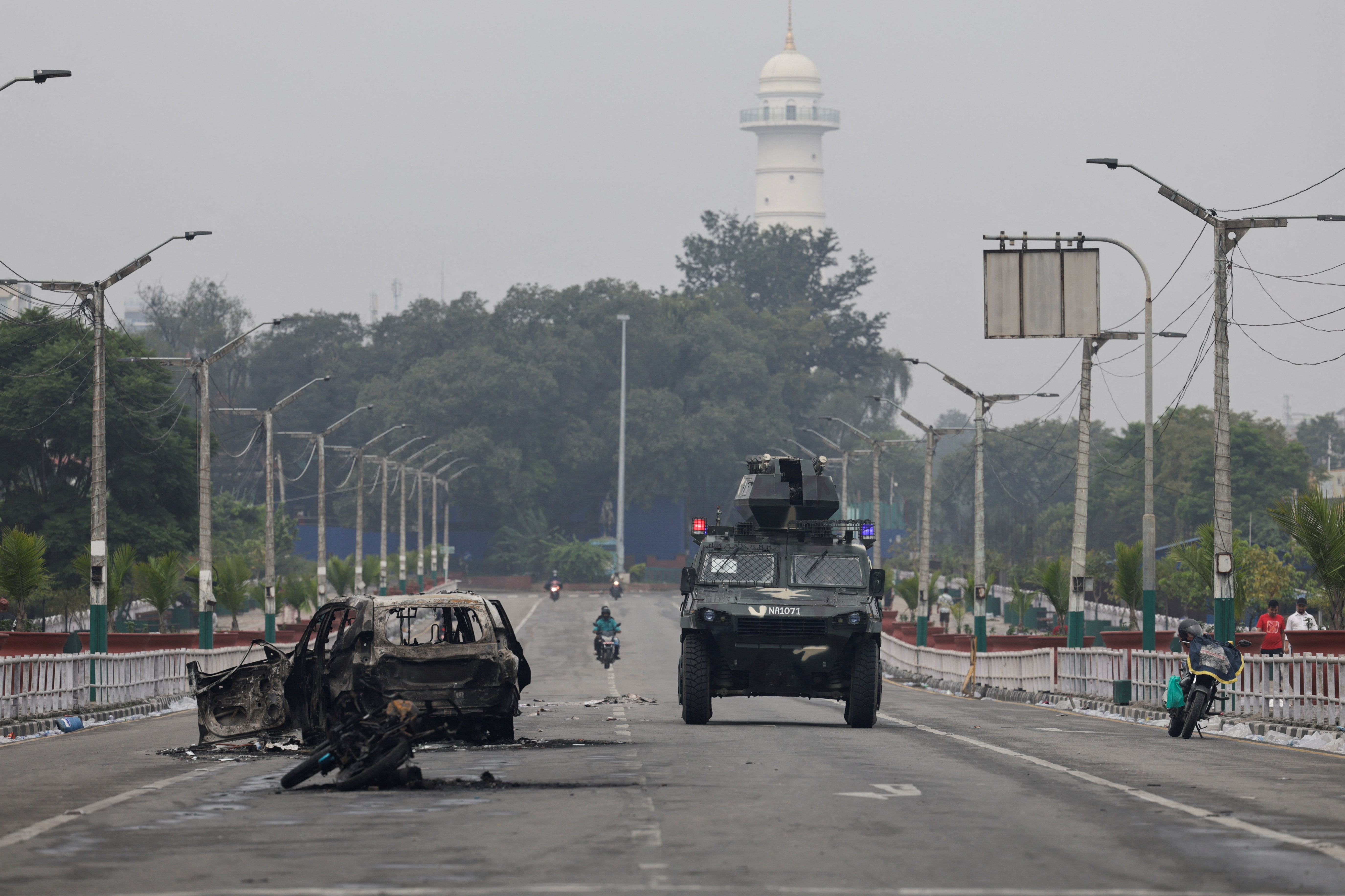 A military vehicle moves past a burnt vehicle on a road near the Singha Durbar office complex that houses the Prime Minister's office