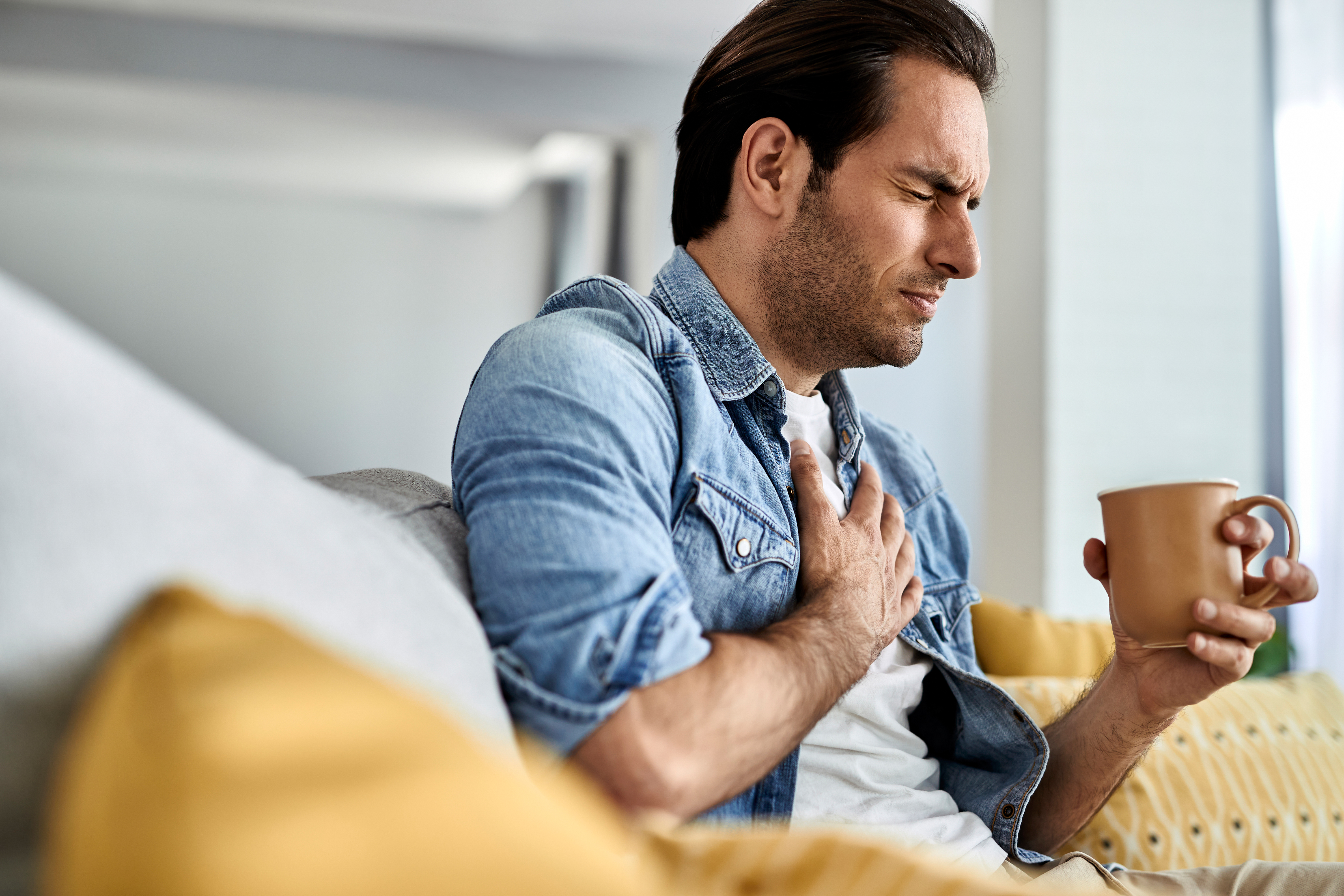 Young man feeling sick and holding his chest in pain while drinking tea in the living room.