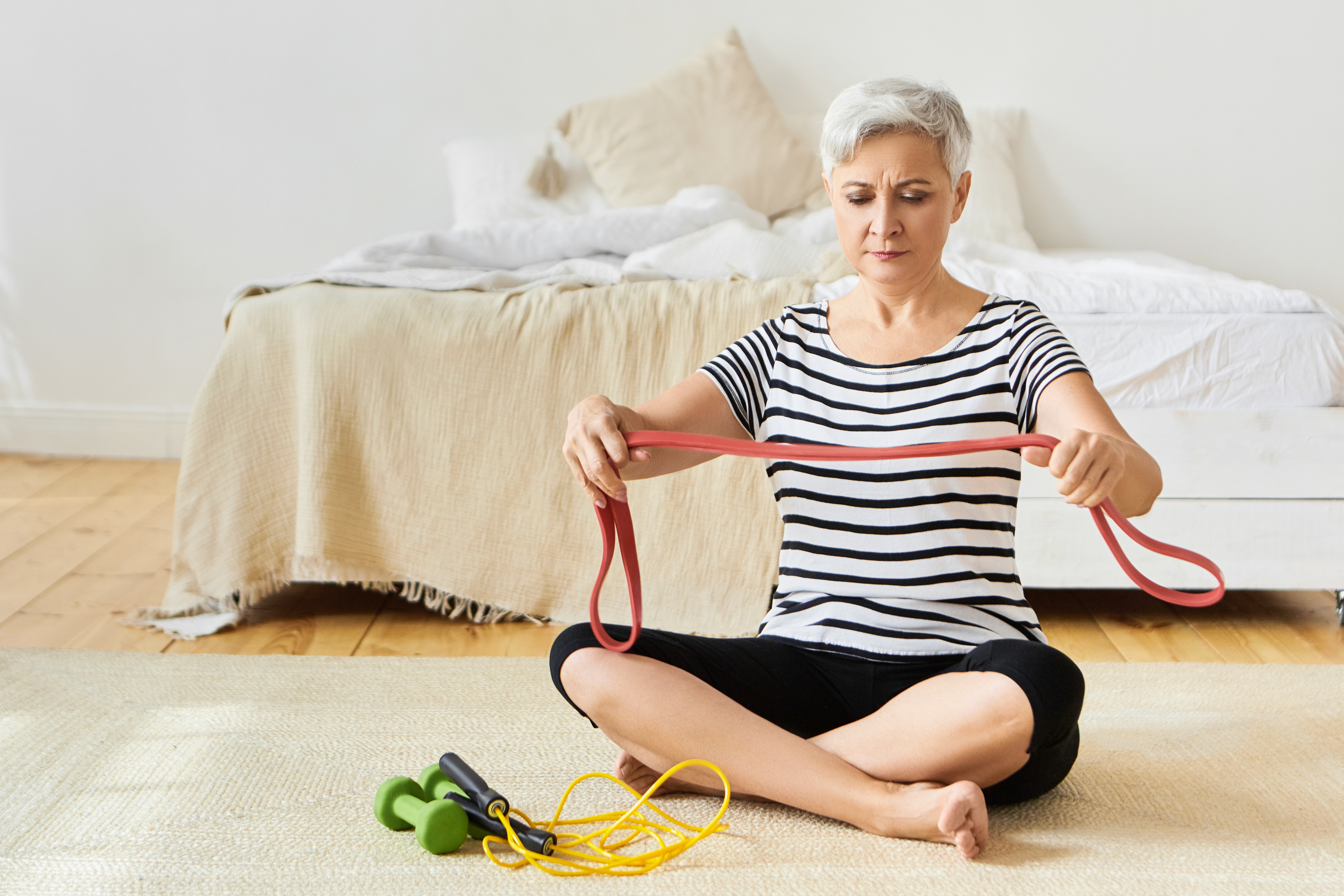 Beautiful concentrated gray haired female pensioner doing exercises for arm muscles using elastic band, sitting on floor with skipping rope and dumbbells. Age, mature people and active lifestyle