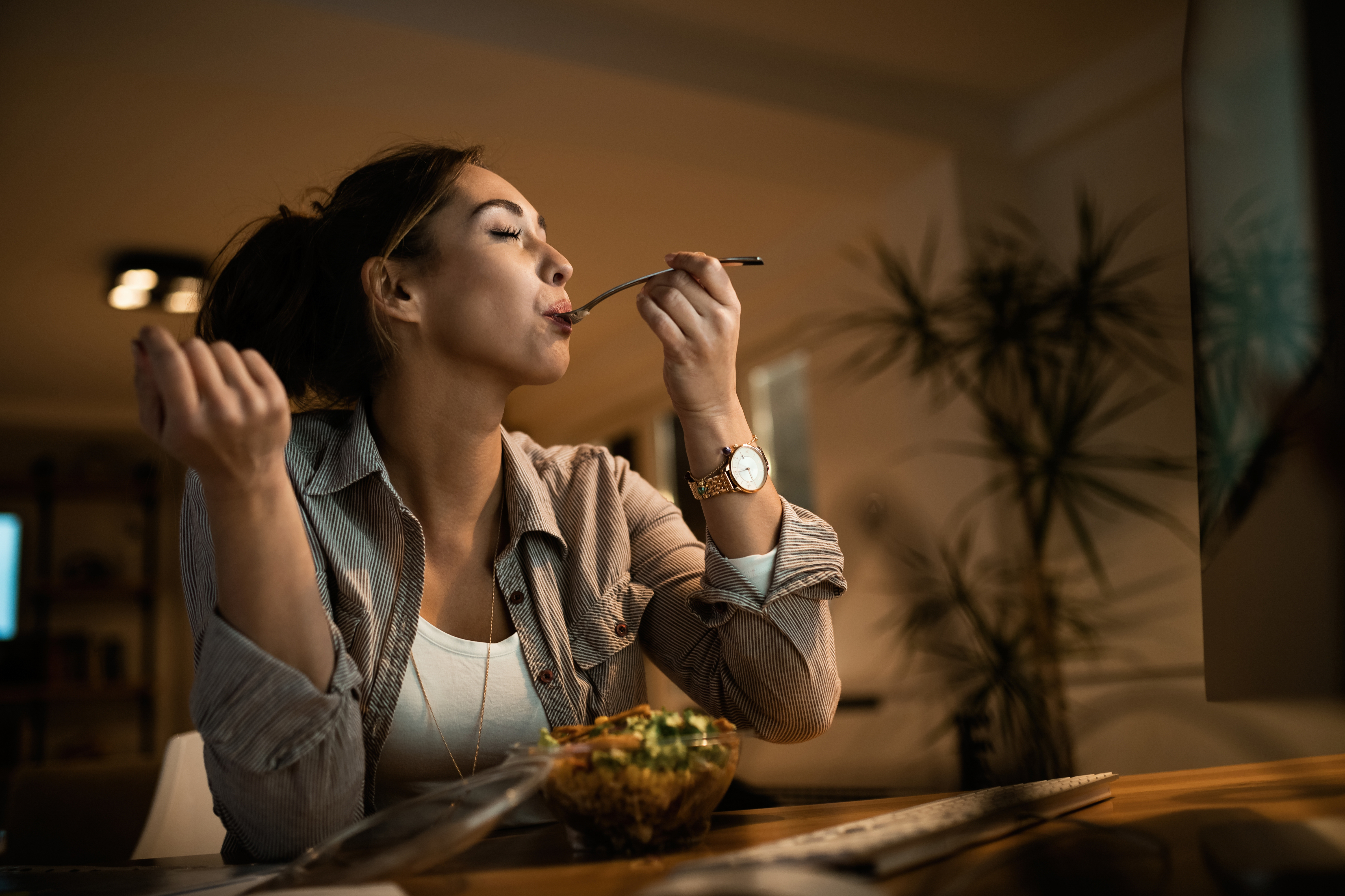 Low angle view of young woman eating salad with her eyes closed while using computer at night at home.