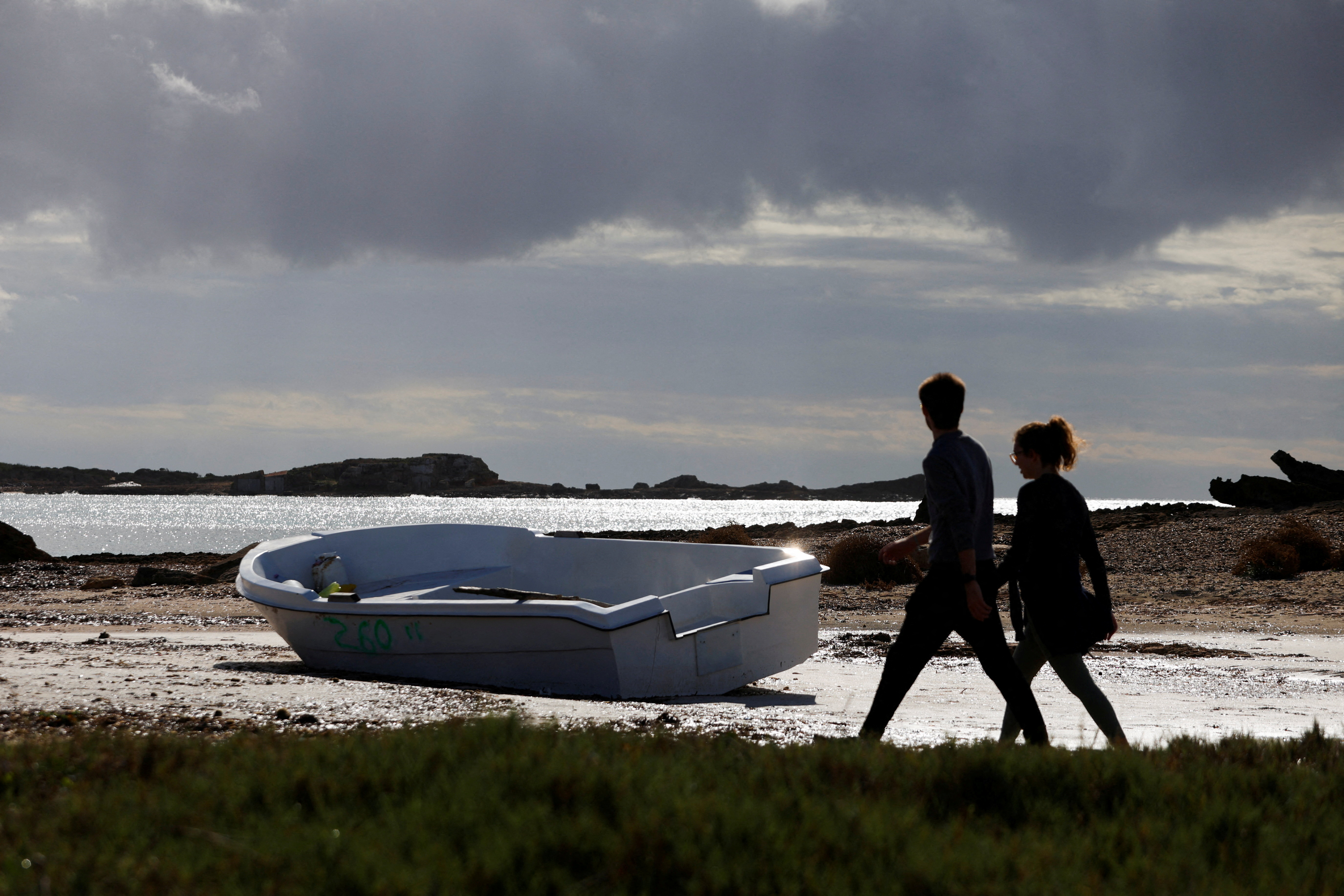 People walk next to a discarded boat used by migrants to reach Mallorca rests on Es Caragol beach in the south of the Balearic island, in Santanyi, Mallorca, Spain