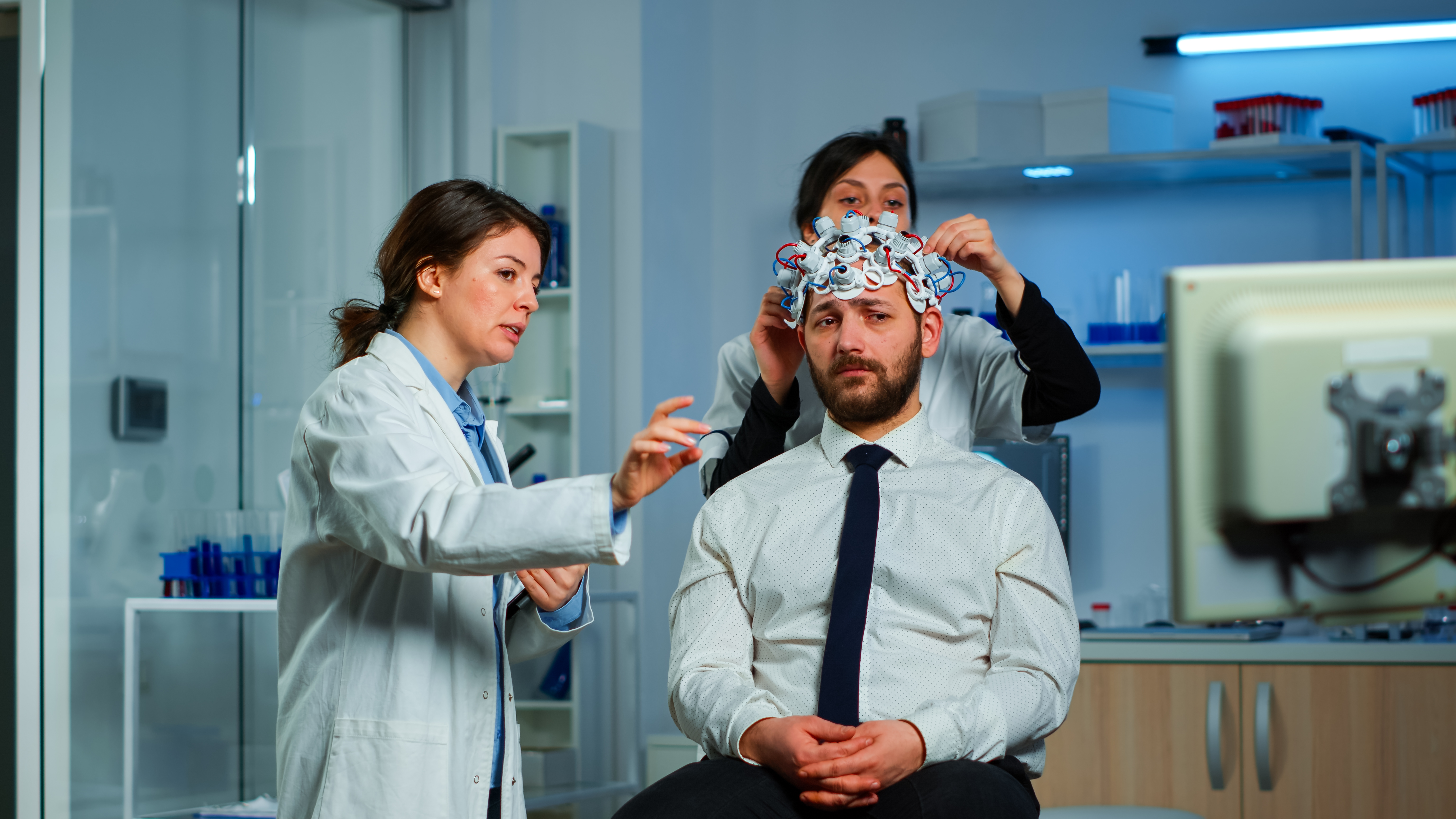 Neurological researchers explaning treatment result pointing on monitor while medical scientist adjusting brainwave scanning headset preparing for brain scan analysing electrical activity
