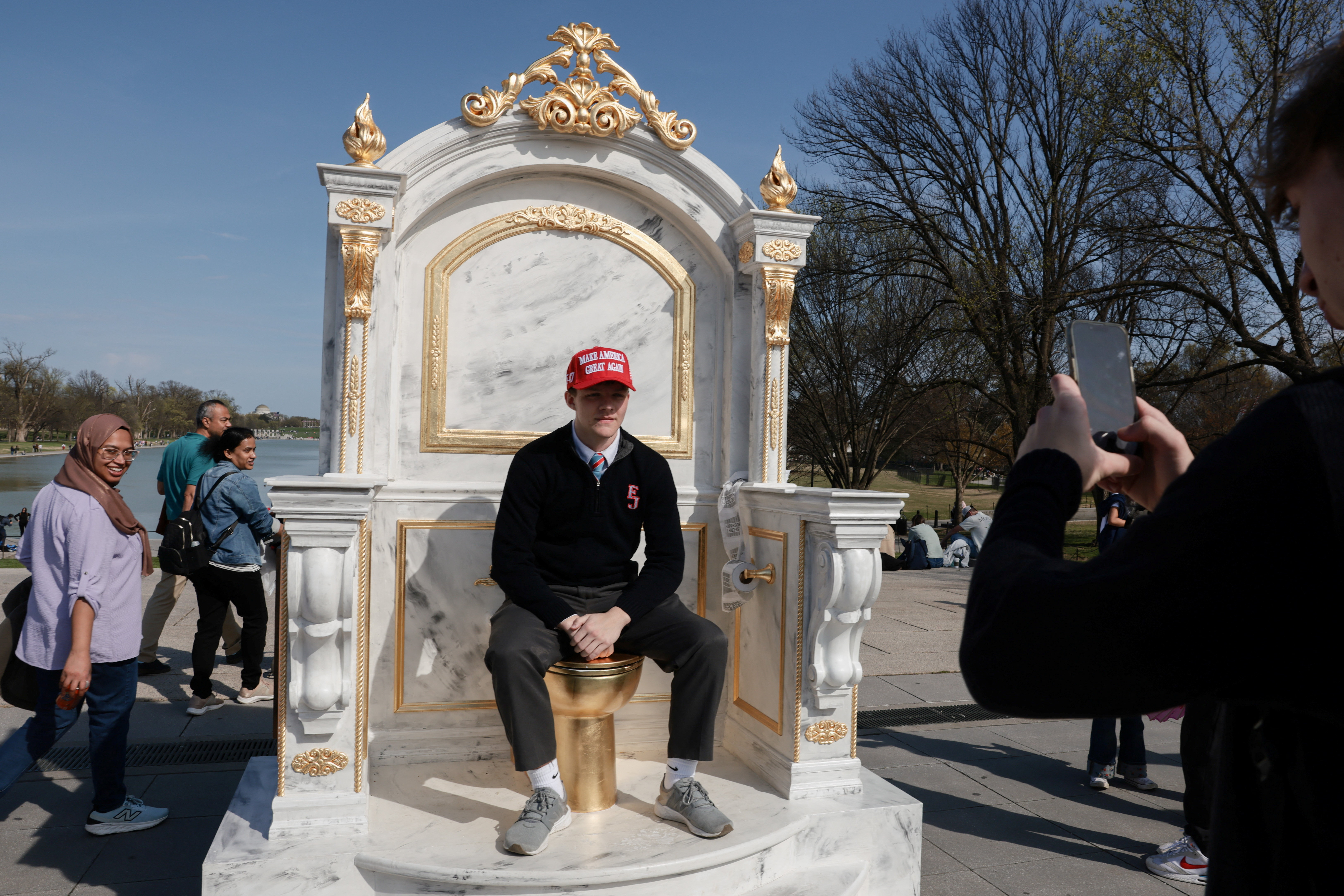 A person wearing a MAGA hat sits on a statue featuring a large golden toilet, titled "A Throne Fit for a King," as it is displayed near the Lincoln Memorial on the National Mall in Washington, D.C., U.S., March 30, 2026.
