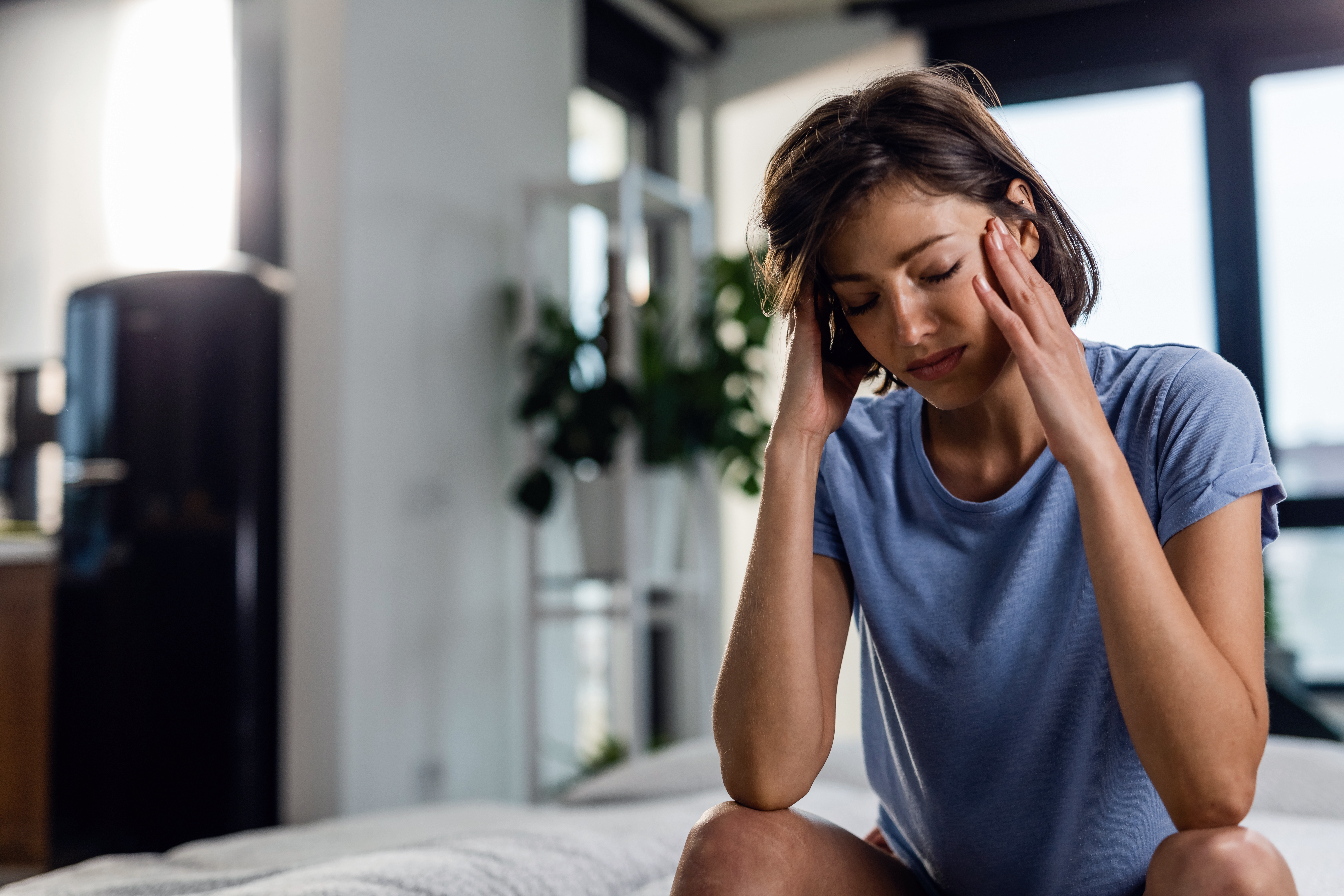 Sad woman suffering from headache and sitting on the bed with eyes closed while holding her head in pain.