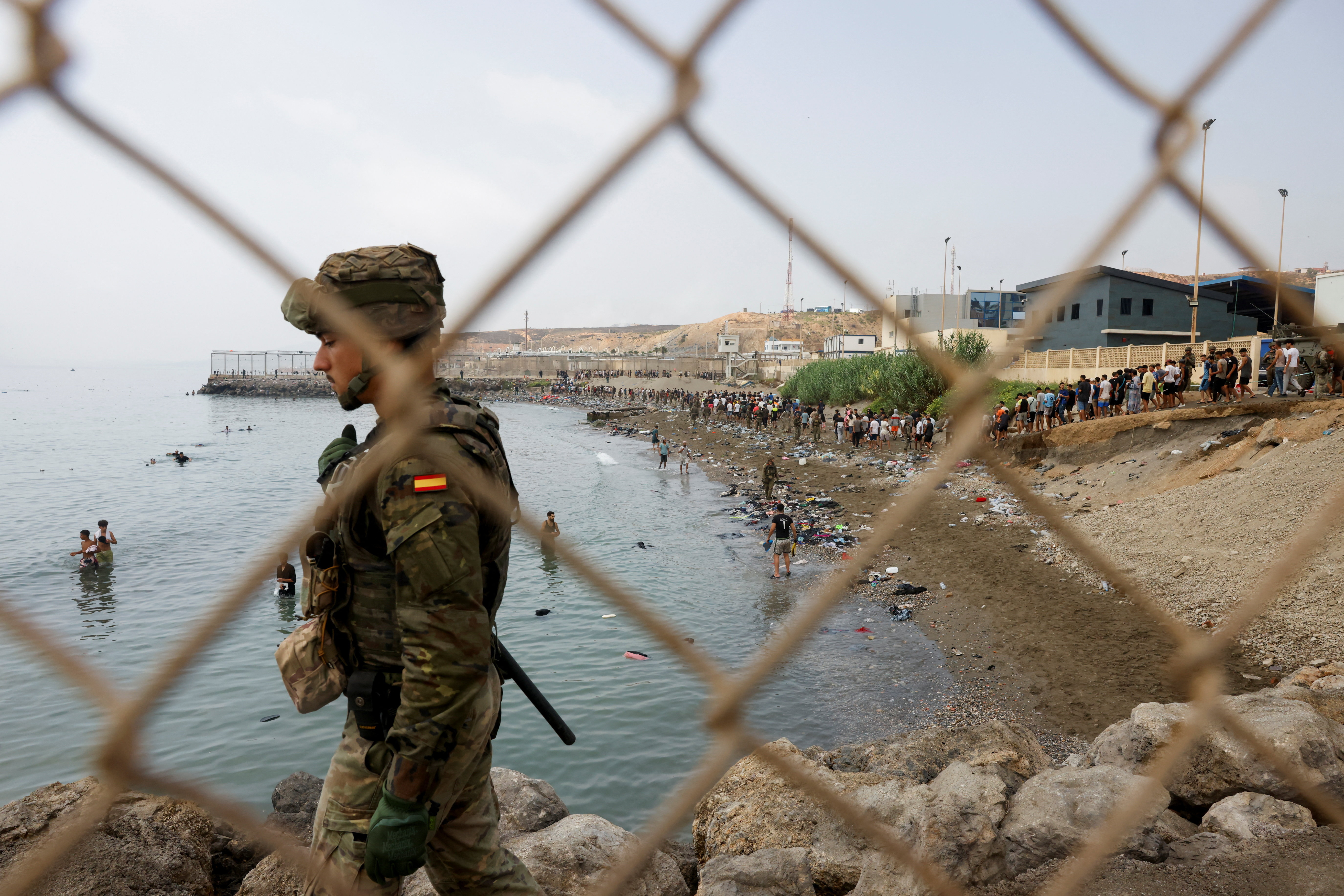 A member of the Spanish military keeps watch by a fence, as people waddle through water attempting to enter Spain, and in the background people who just entered Spain walk back to return to Morocco