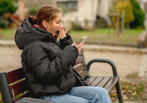 Girl Yawning While Sitting With Smartphone On A Bench In Spring