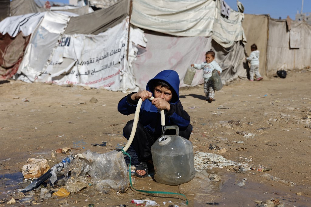 A displaced Palestinian child fills a water container from a hose pipe, as a toddler runs over with empty containers from a family tent shelter, set-up with hundreds of other shelters, in Gaza City