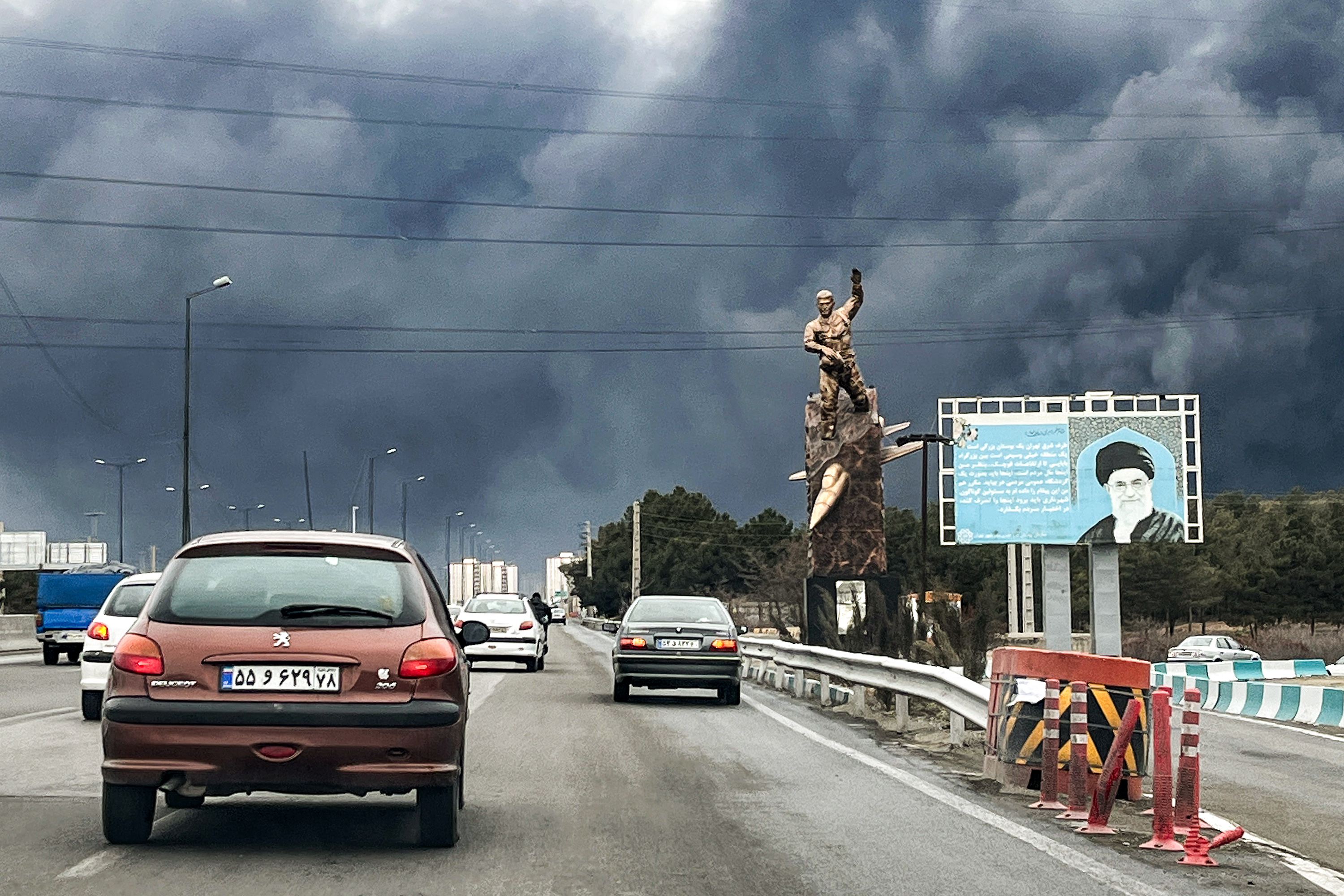 Vehicles move along a highway past a war memorial statue and a billboard depicting Iran's late supreme leader Ayatollah Ali Khamenei