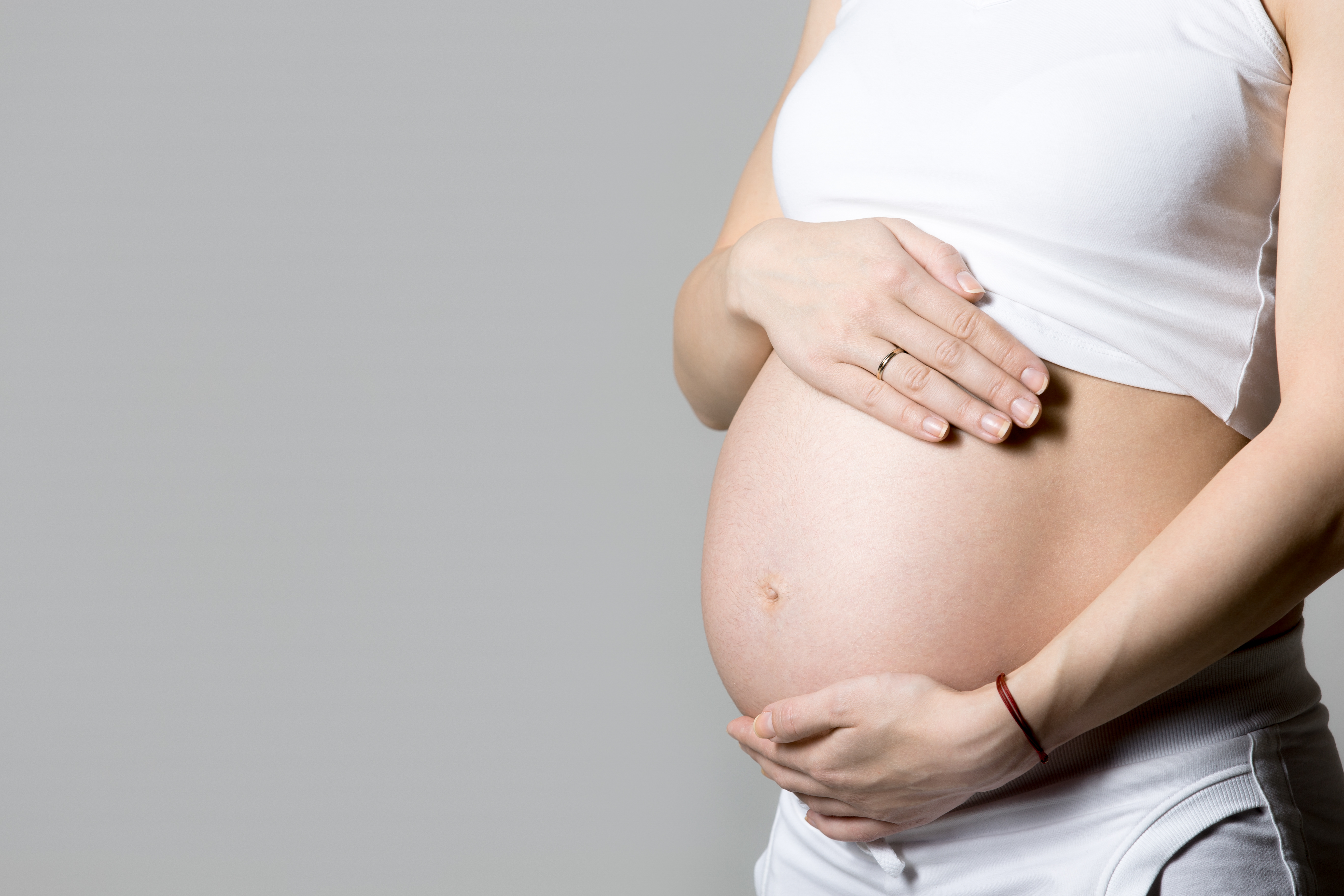 Young pregnant model in tank top touching her belly, gray background, studio, copy space, close-up
