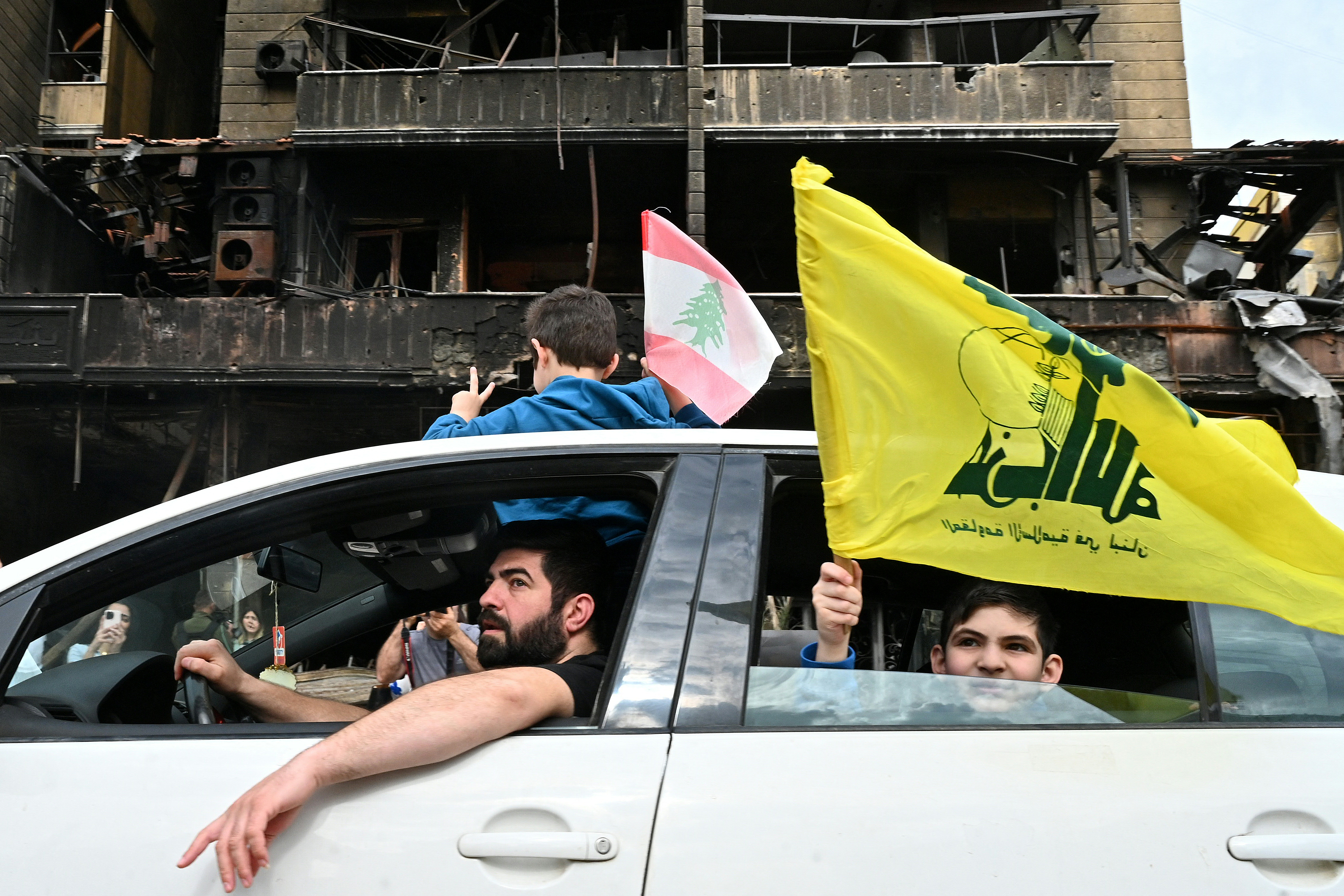 Boys wave Lebanese and Hezbollah flags while going past a heavily damaged building as displaced people return to their neighbourhood in Beirut's southern suburbs after a 10-day ceasefire with Israel came into effect