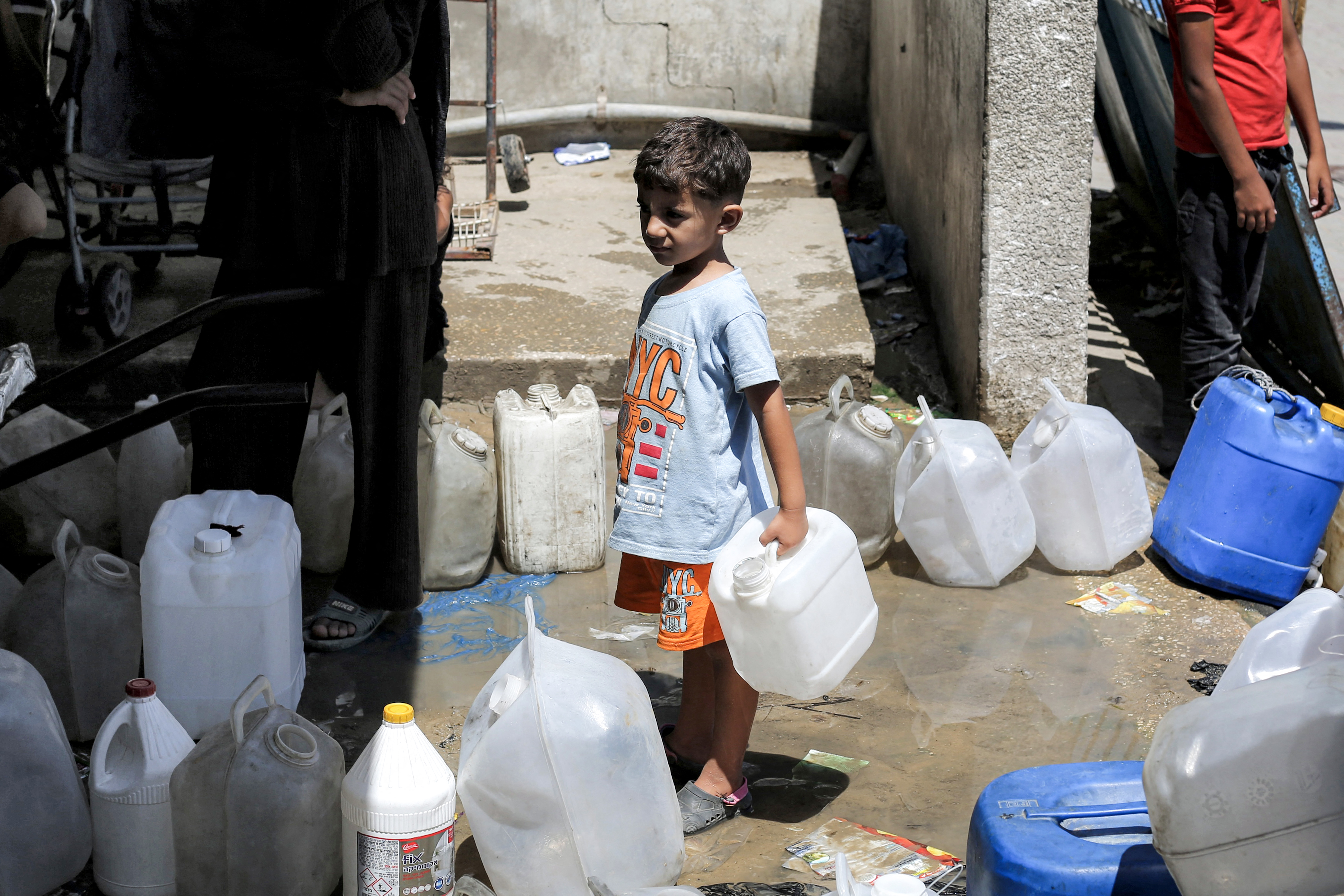 A boy stands next to other people queueing to fill up their containers from a cistern at the Maghazi camp for Palestinian refugees in the central Gaza Strip on July 26, 2026.